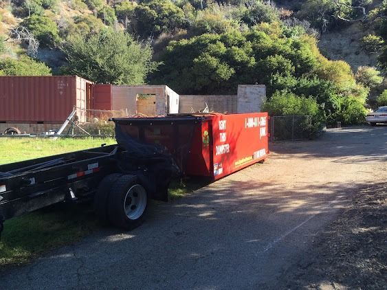Red dumpster on a trailer, parked on a paved road. Containers and hillside in the background.