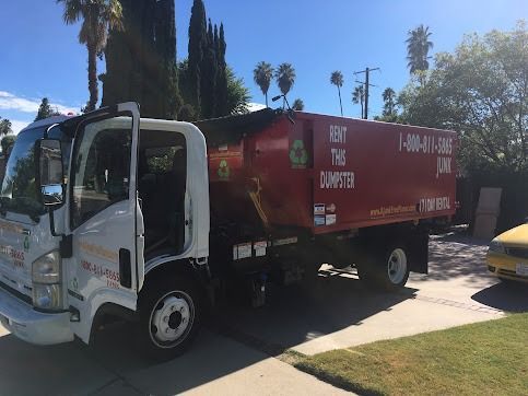 White and red roll-off dumpster truck parked on a driveway with a tree-lined background. The door is open.