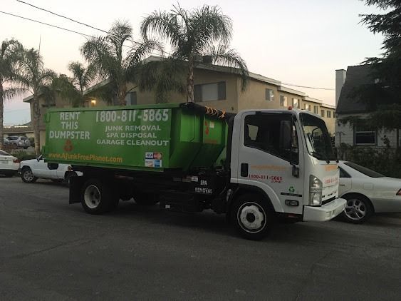 Green dumpster truck parked on a street in front of an apartment building.