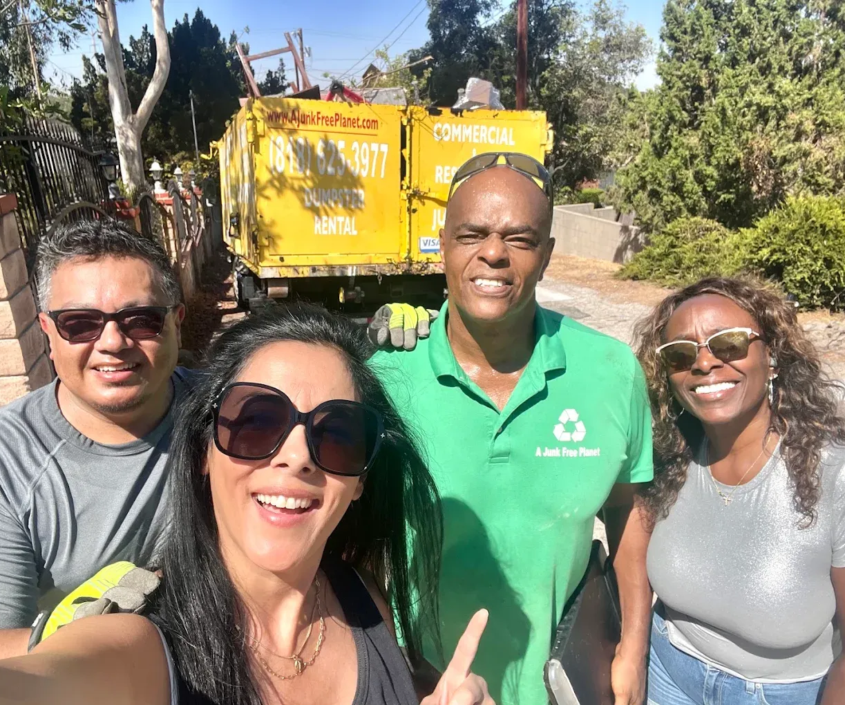 Four people stand in front of a yellow dumpster, smiling. One points; setting appears outdoors.