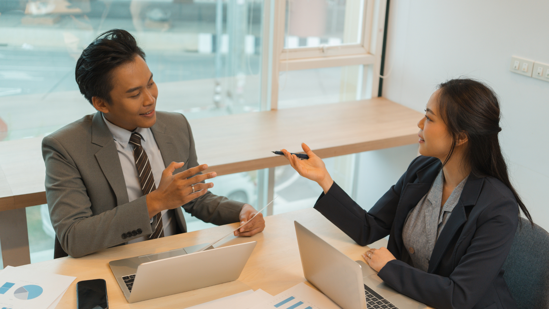 Two people in business attire at a desk, laptops open, conversing.