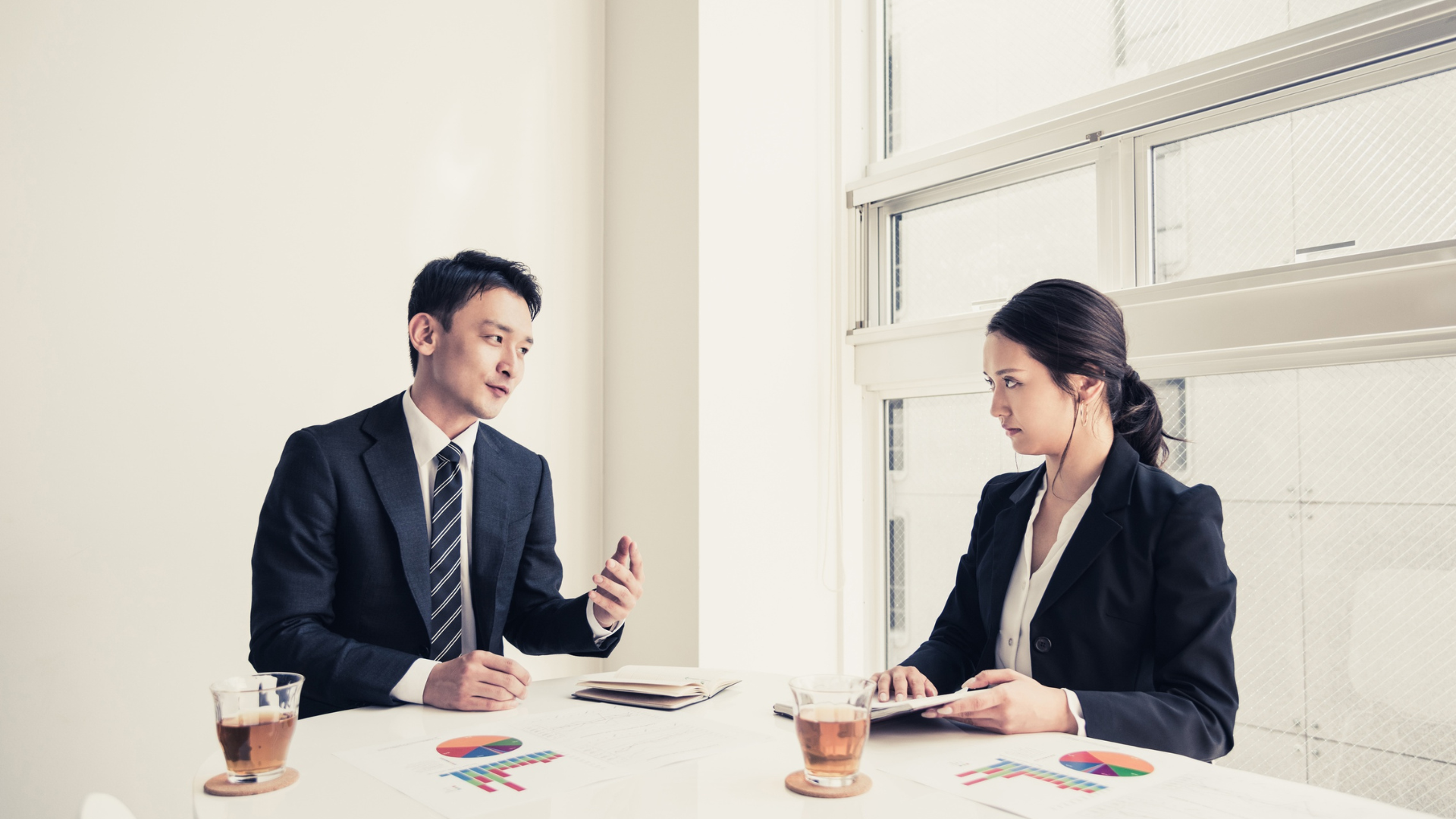 Man and woman in business suits at a table; man gestures while talking, woman listens intently. White wall, window.