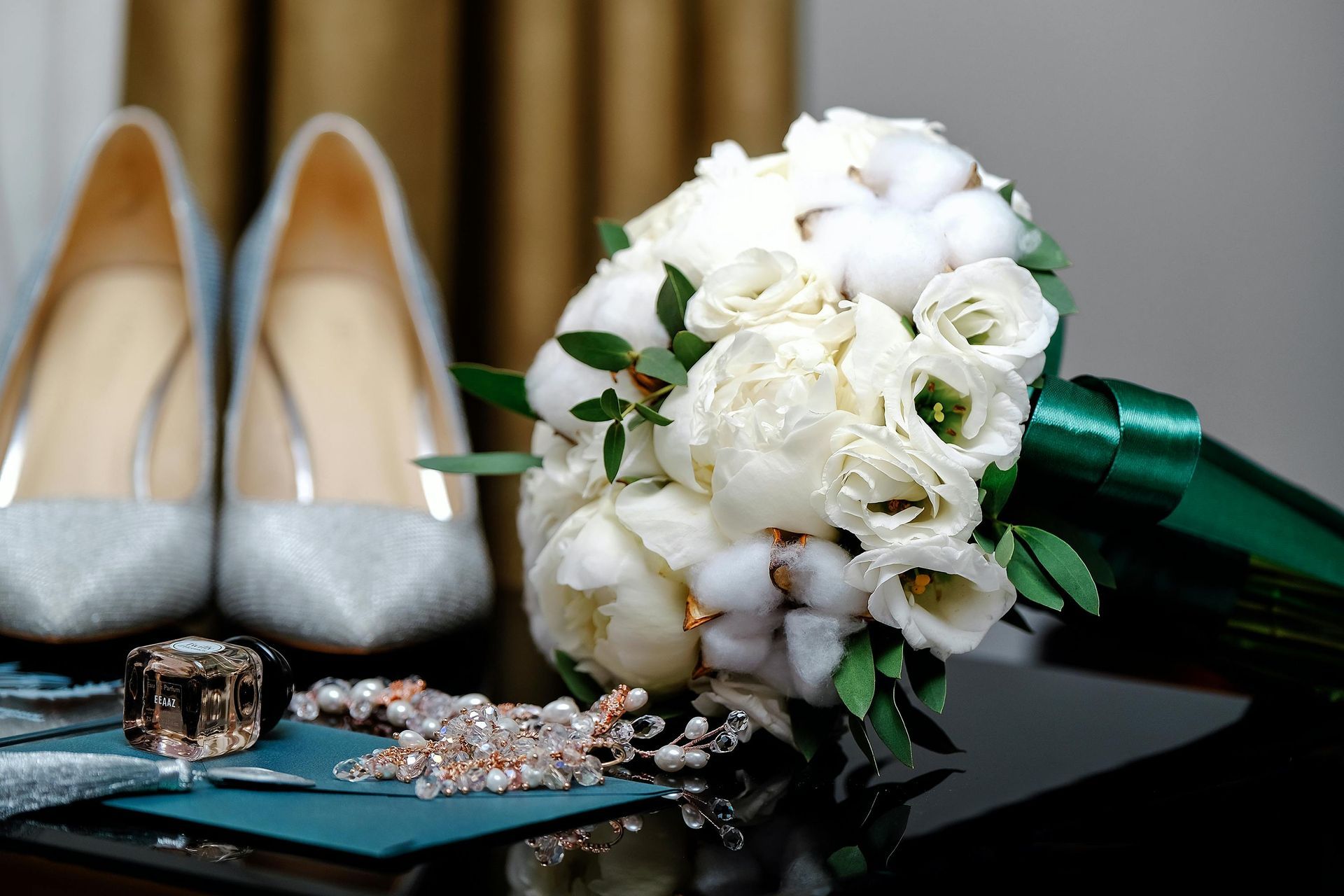 A bridal bouquet of white peonies and cotton bolls with a green ribbon, set beside sparkling heels and jewelry.