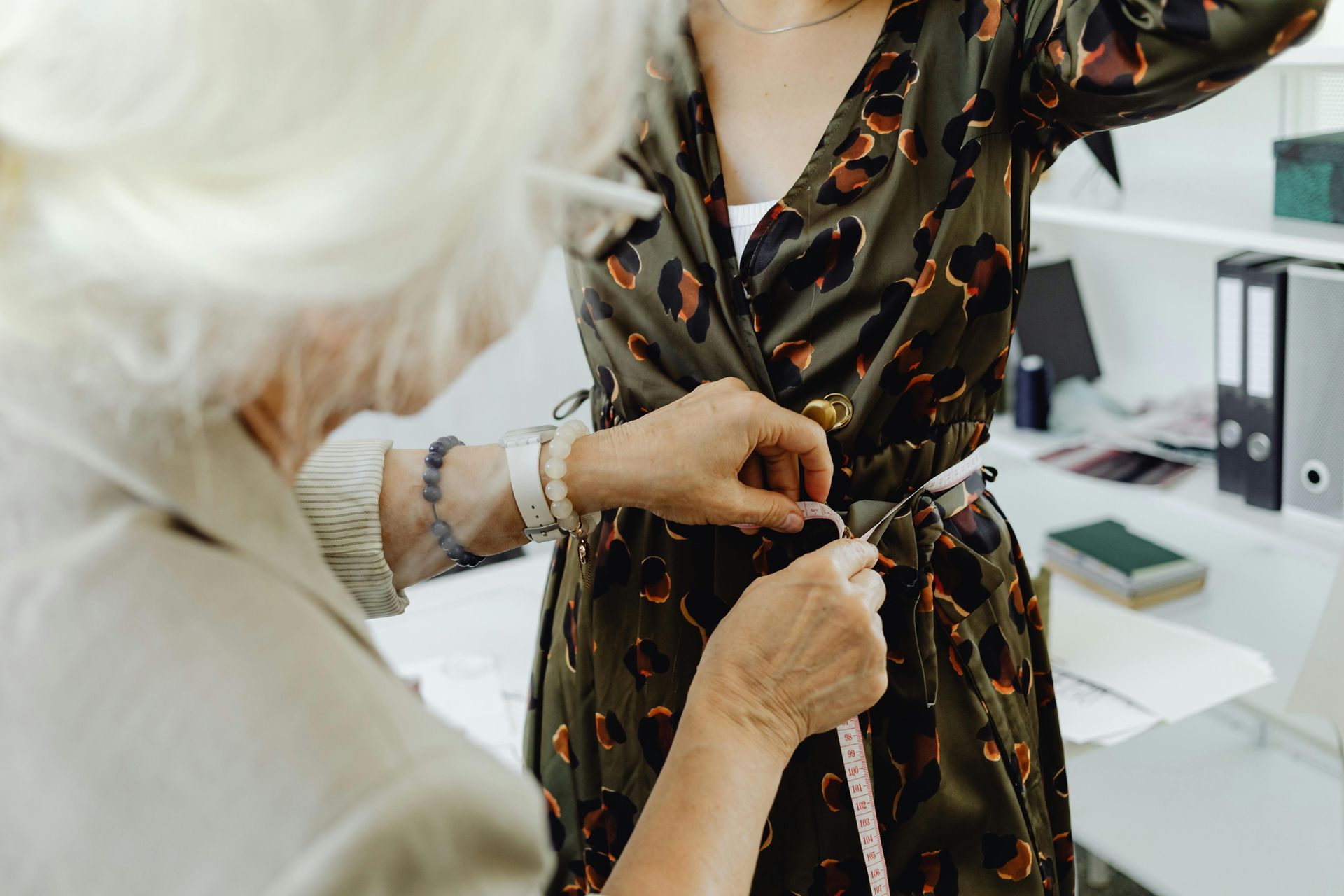 A seamstress measures a dress on a person in a brightly lit room with office supplies on a shelf.