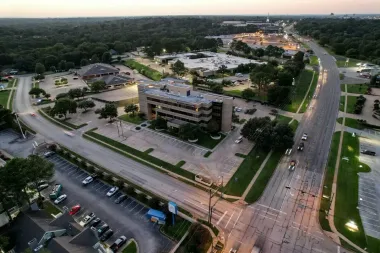 Aerial view of a city intersection with buildings, parking lots, and tree-lined roads at dusk