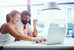 Two coworkers smiling at a laptop in a bright office, one pointing at the screen