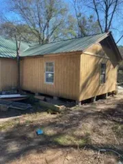 A small wooden house with a green roof is sitting in the middle of a dirt field.