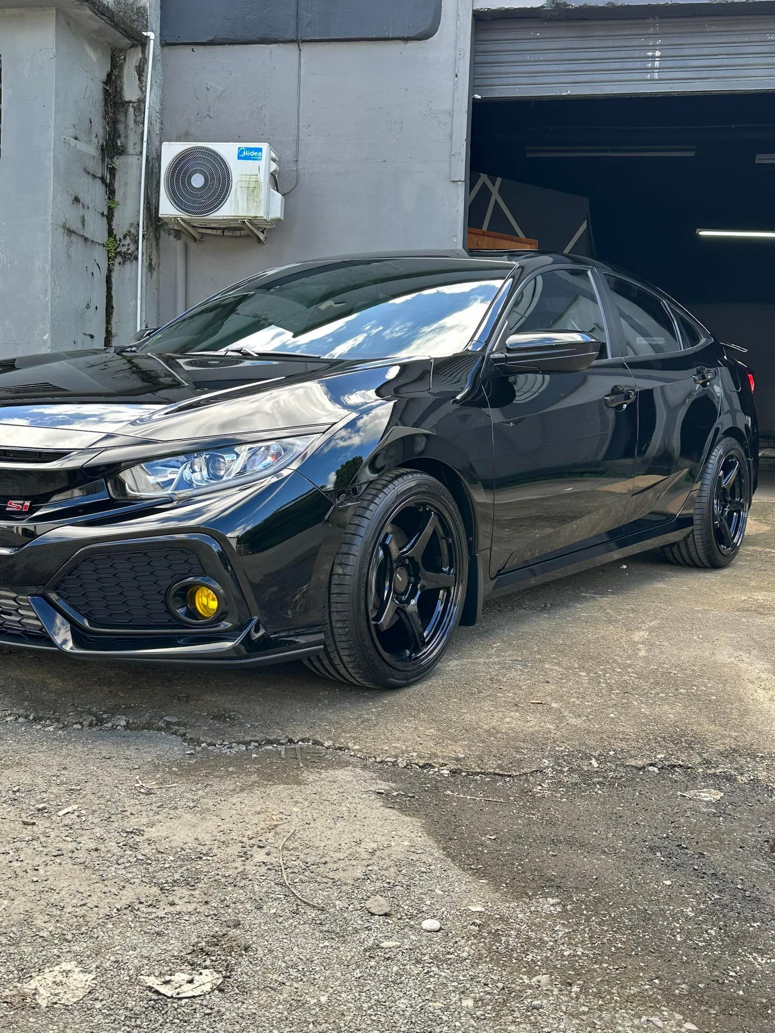 Black Honda Civic car parked outside, yellow fog lights, black wheels, shiny paint.