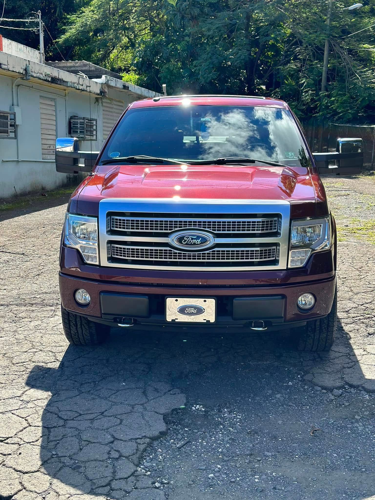 Red Ford F-150 pickup truck parked on a paved surface. Bright sunlight and leafy trees are in the background.