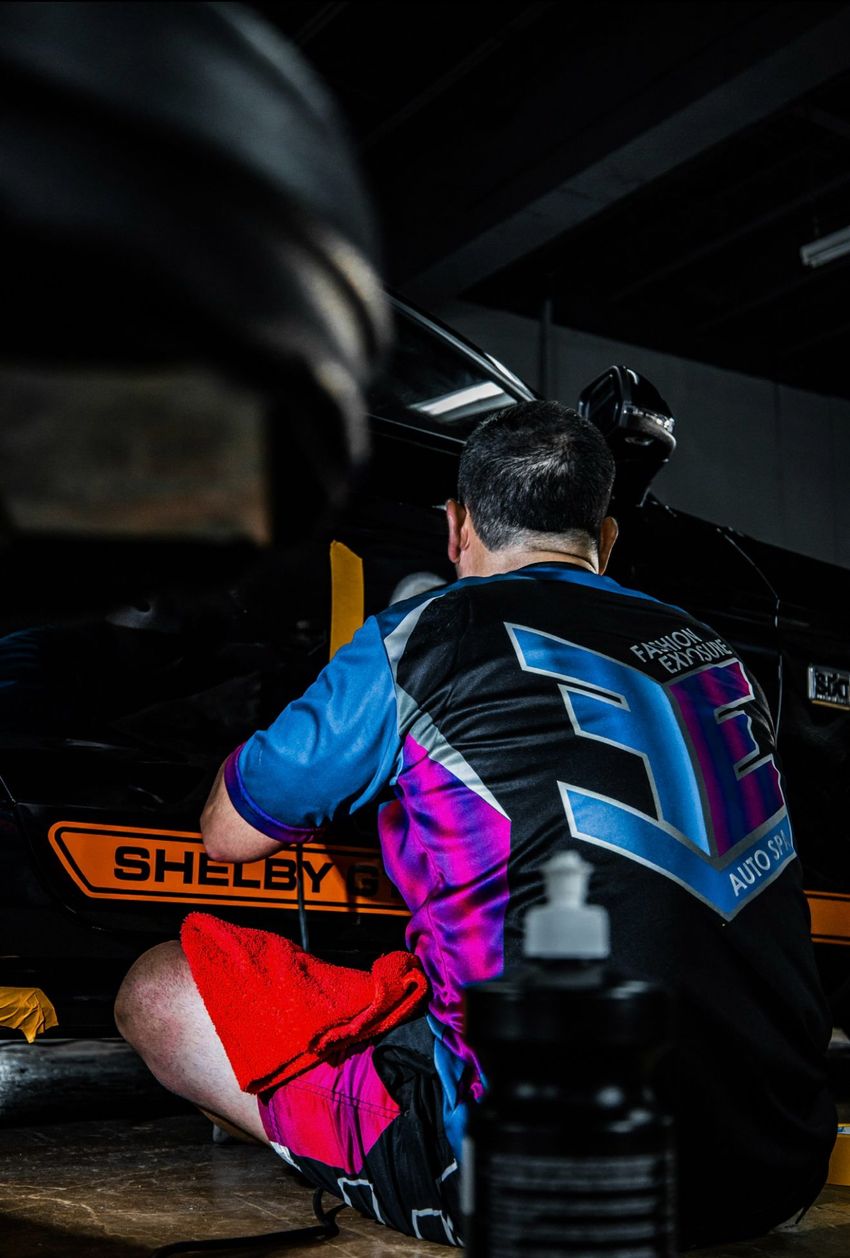 Five men in black shirts pose in front of a turquoise car and a banner with the word