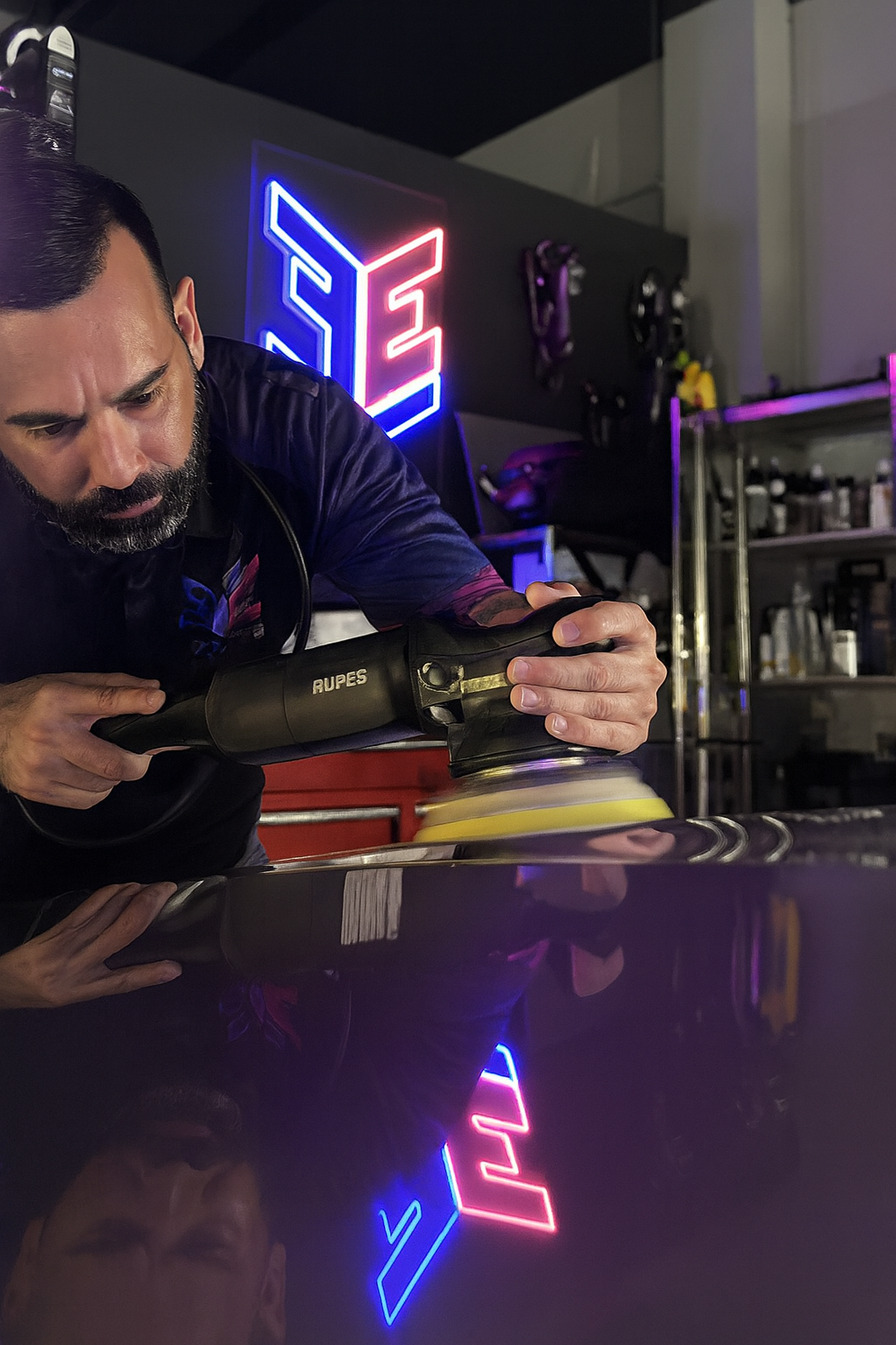 Man polishing a red car bumper, kneeling with a buffer in hand. Interior setting.