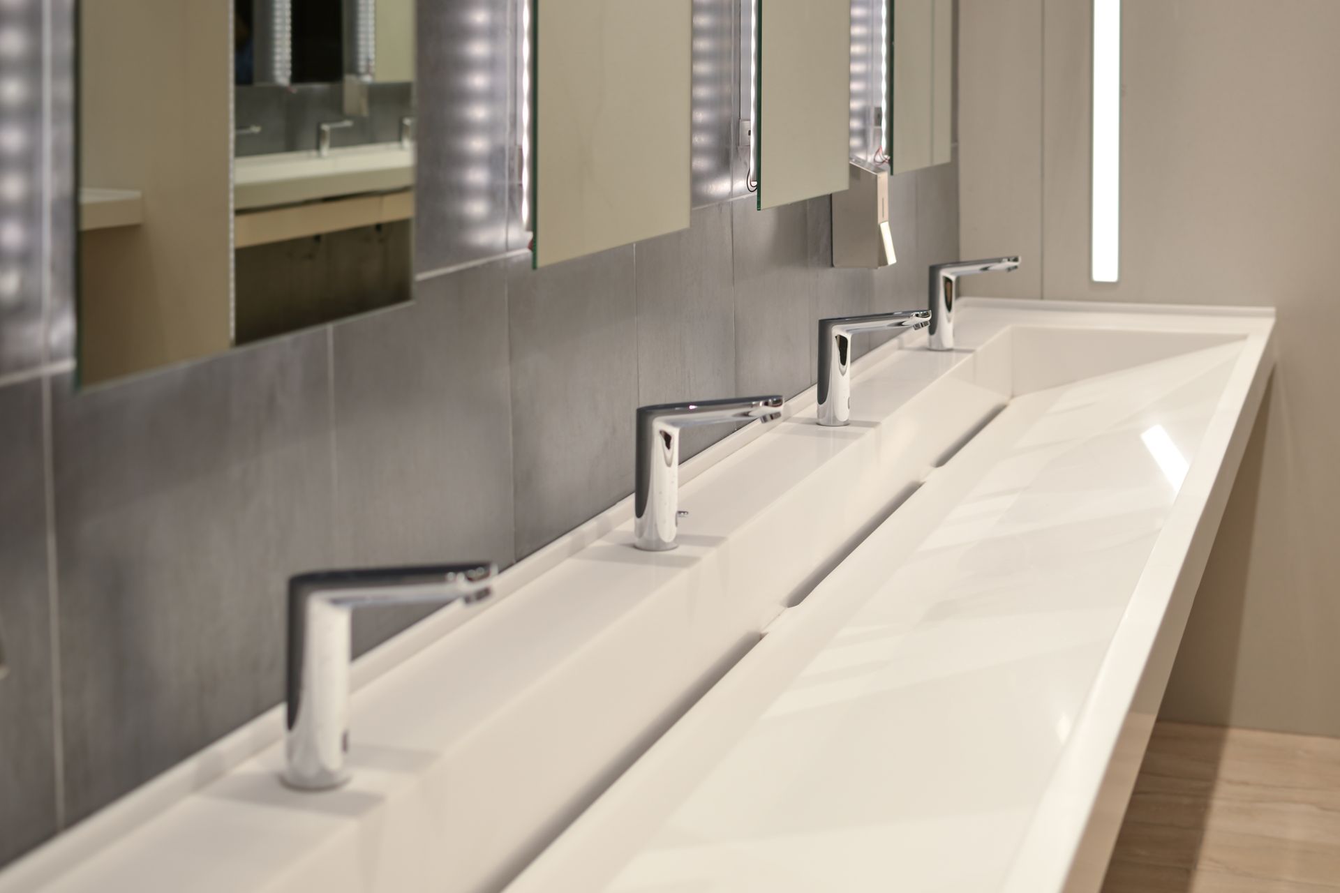 Modern public restroom with a long white sink, chrome faucets, mirrors, and gray tile.
