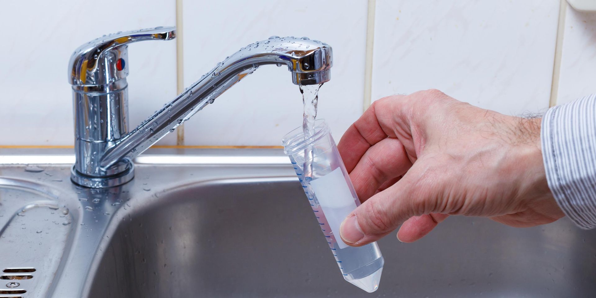 A hand holds a test tube under a faucet, filling it with water in a stainless steel sink.