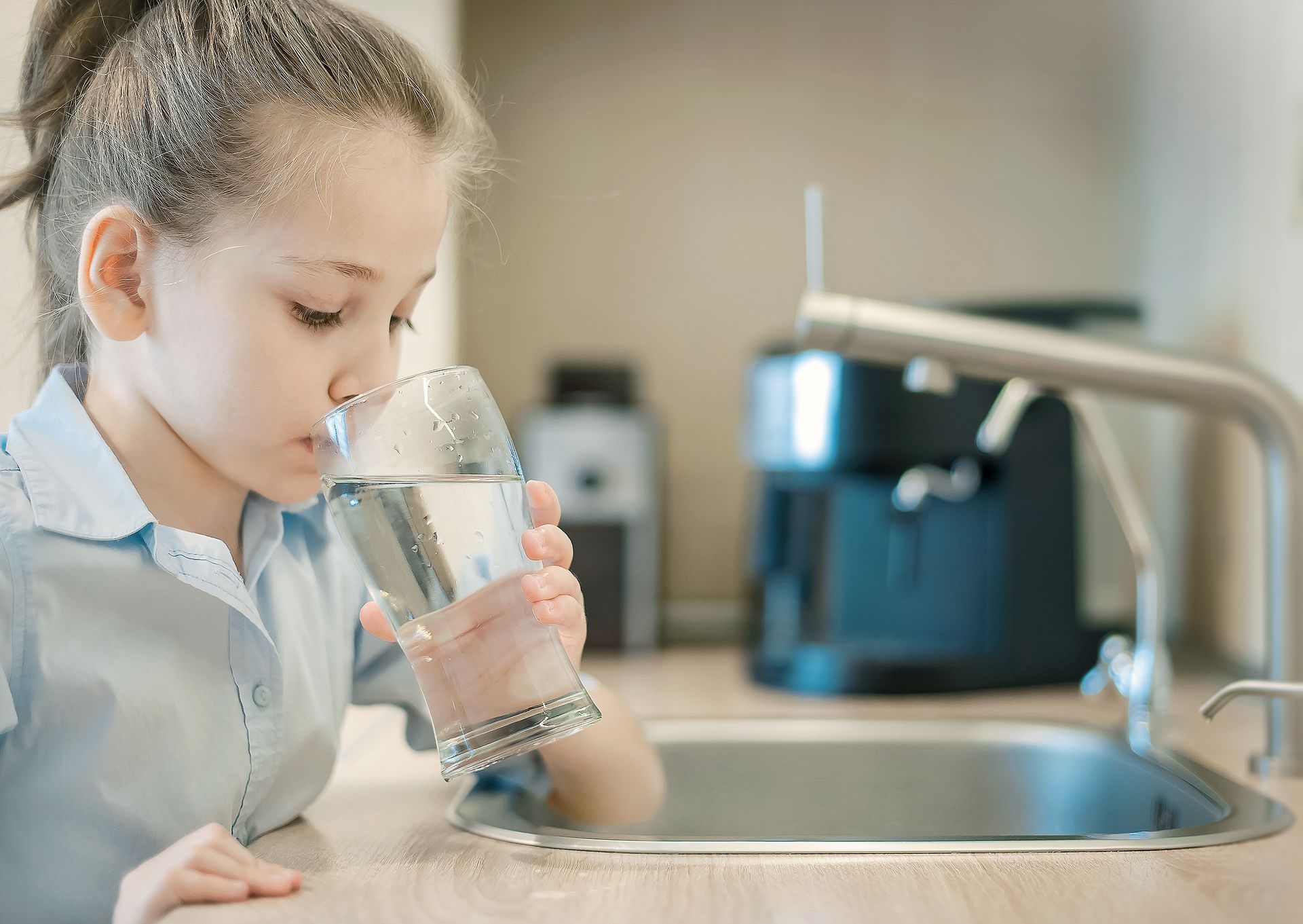 Girl drinking water from a glass in a kitchen, faucet and water filter visible.