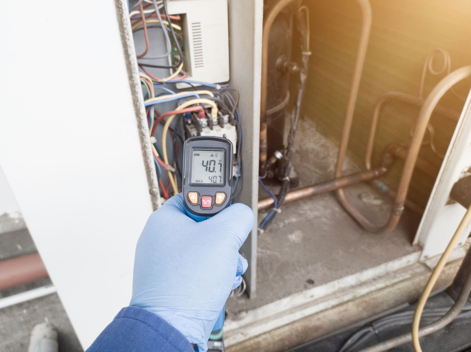 A gloved hand uses an infrared thermometer to measure the temperature of an HVAC unit's electrical components.