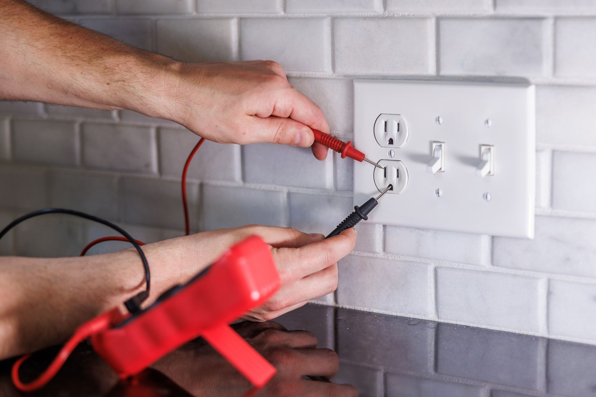 Hands testing electrical outlet with multimeter probes on white tile background.