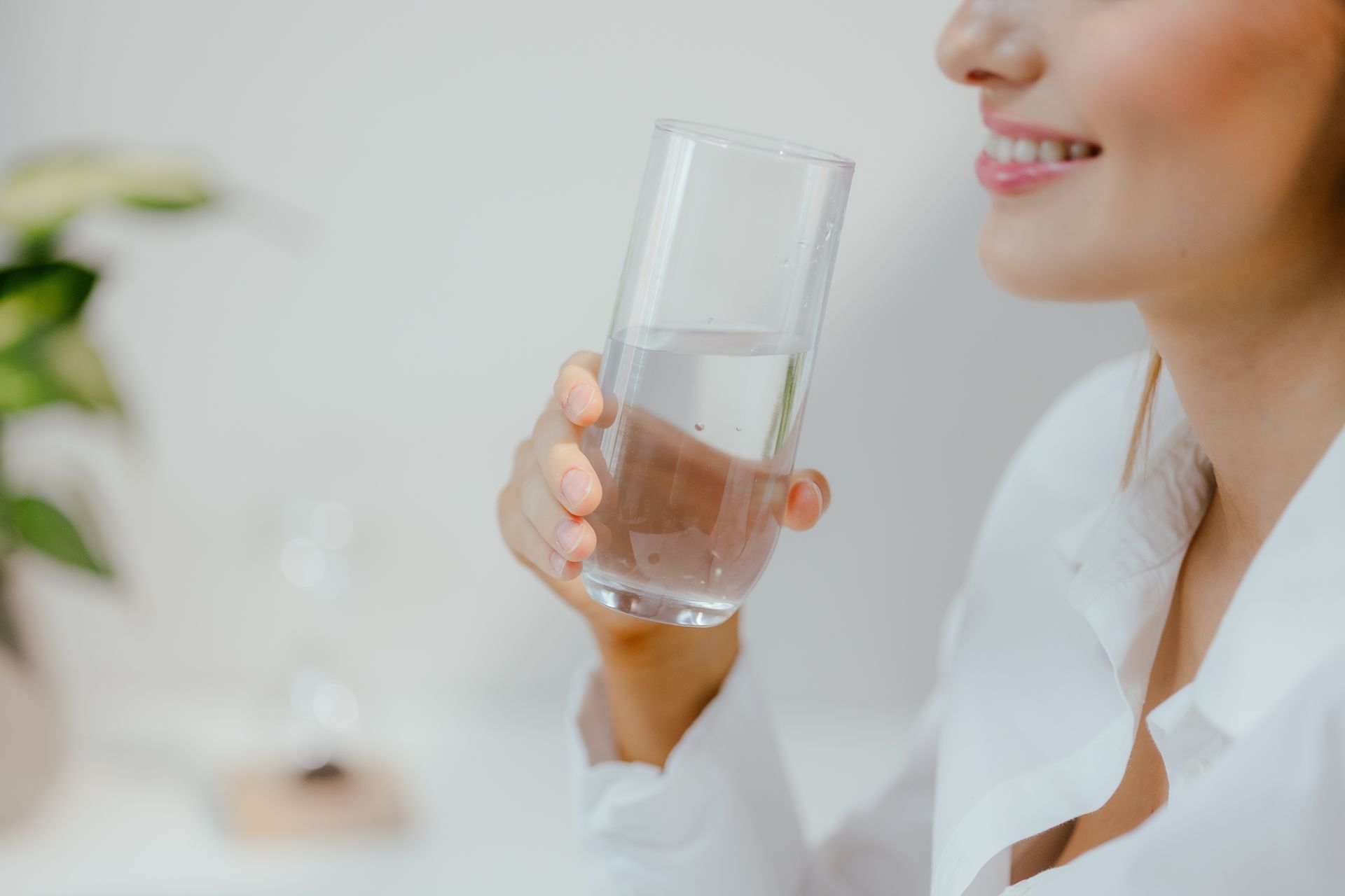 Woman smiling, holding a glass of water, taking a sip, indoors.