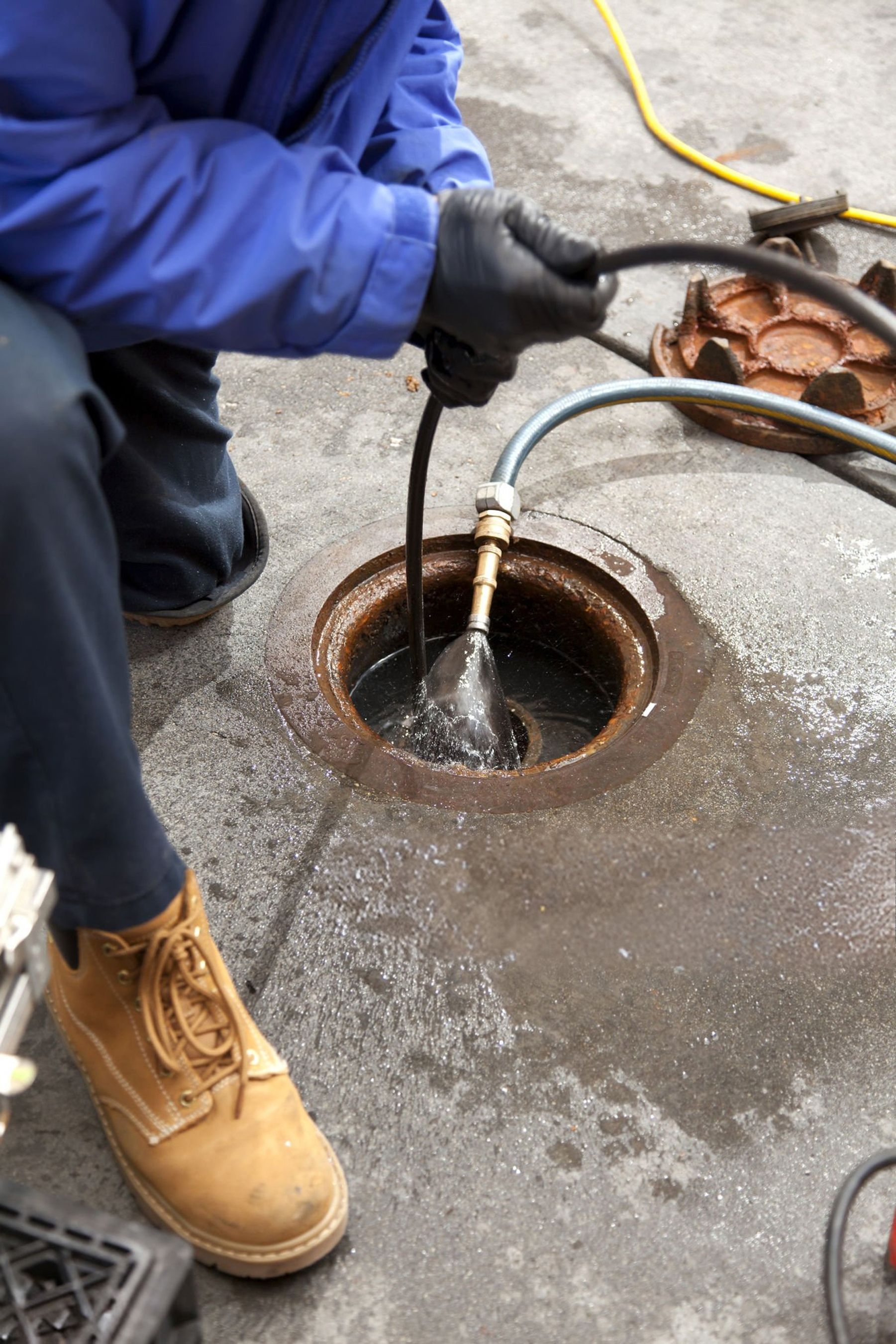 Person in blue jacket and work boots cleaning a dark manhole with a hose on a grey concrete surface.