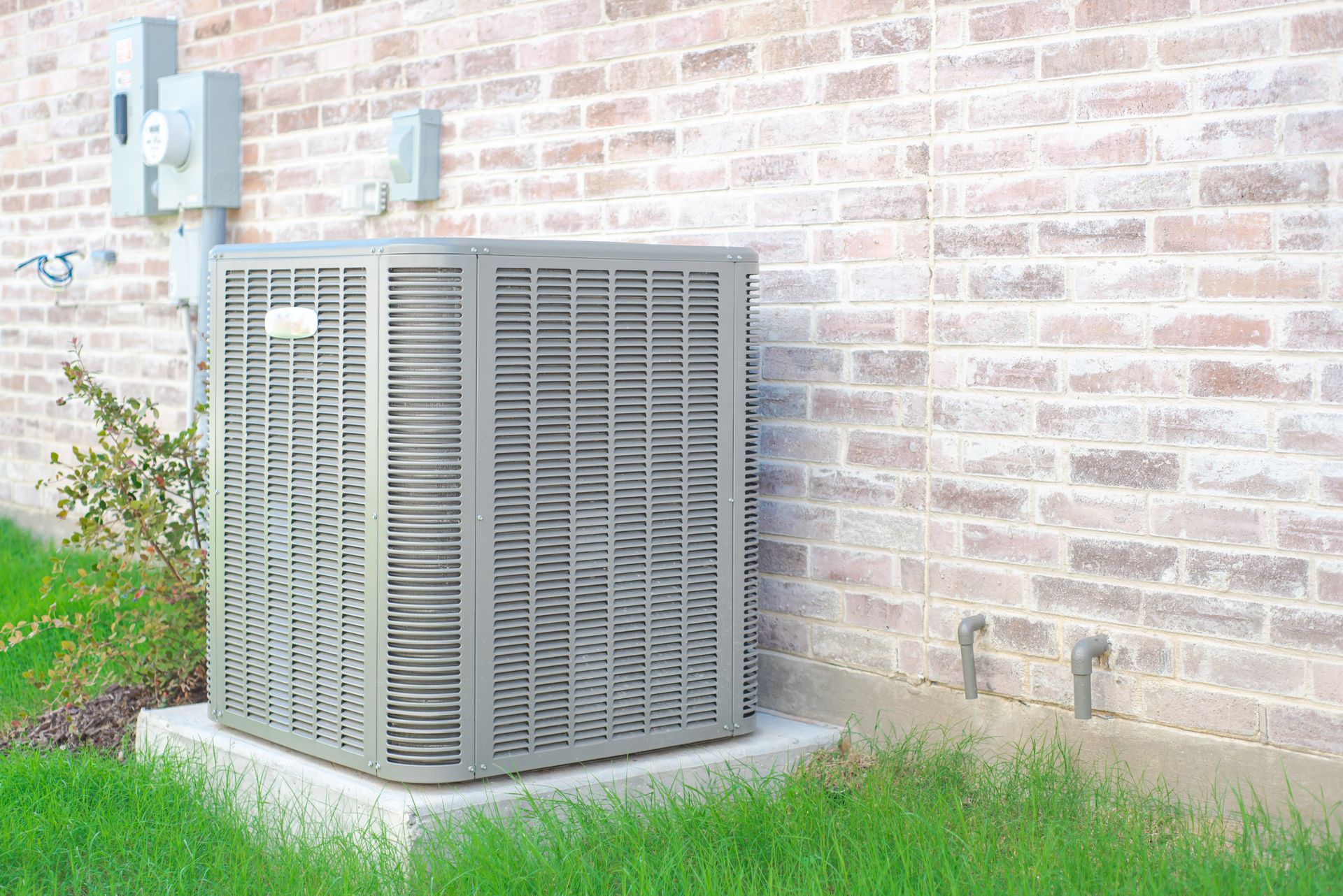 A gray central air conditioning unit sits on a concrete pad against a light brick house wall with electrical equipment.