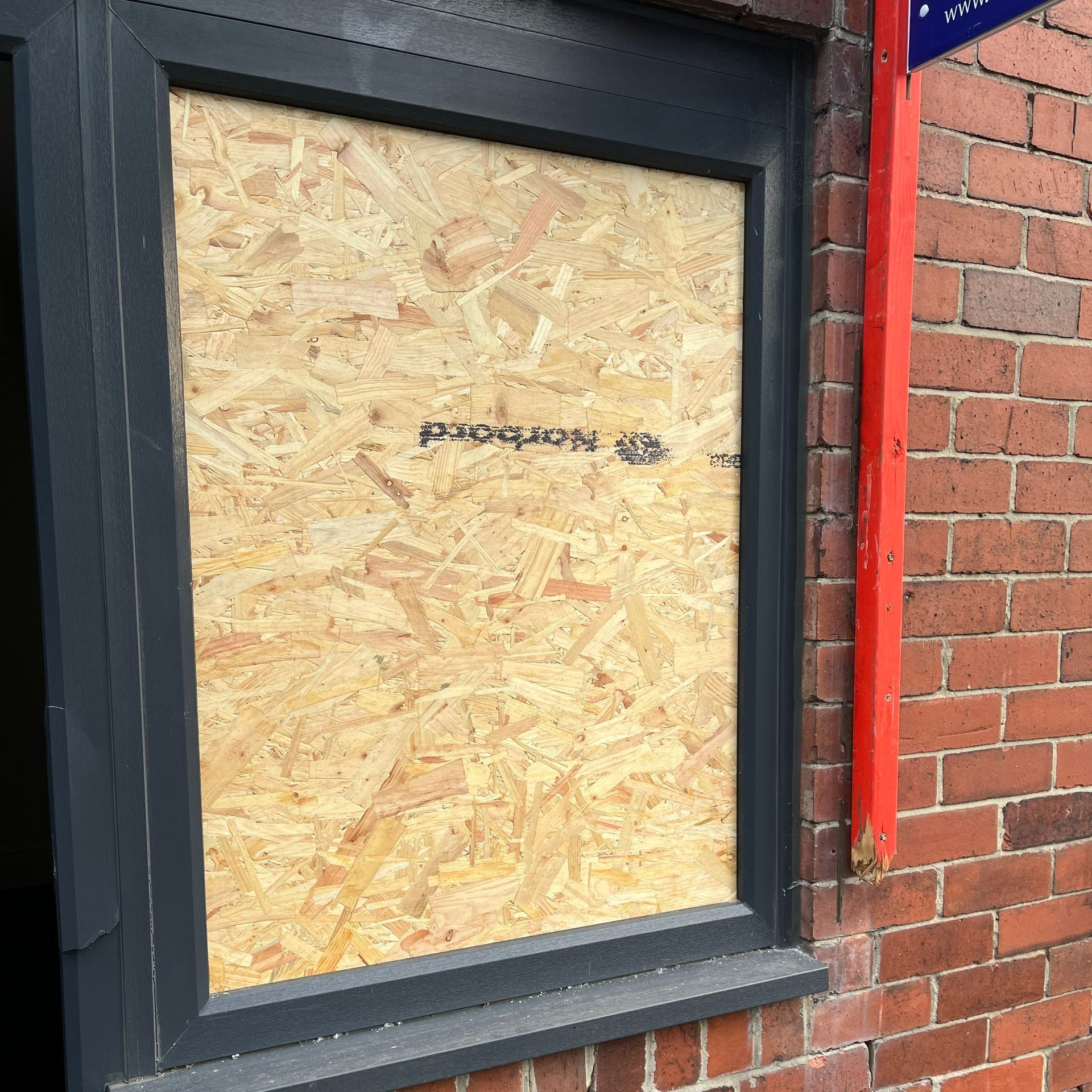Boarded-up window in dark frame on a brick building; red vertical beam to the right.
