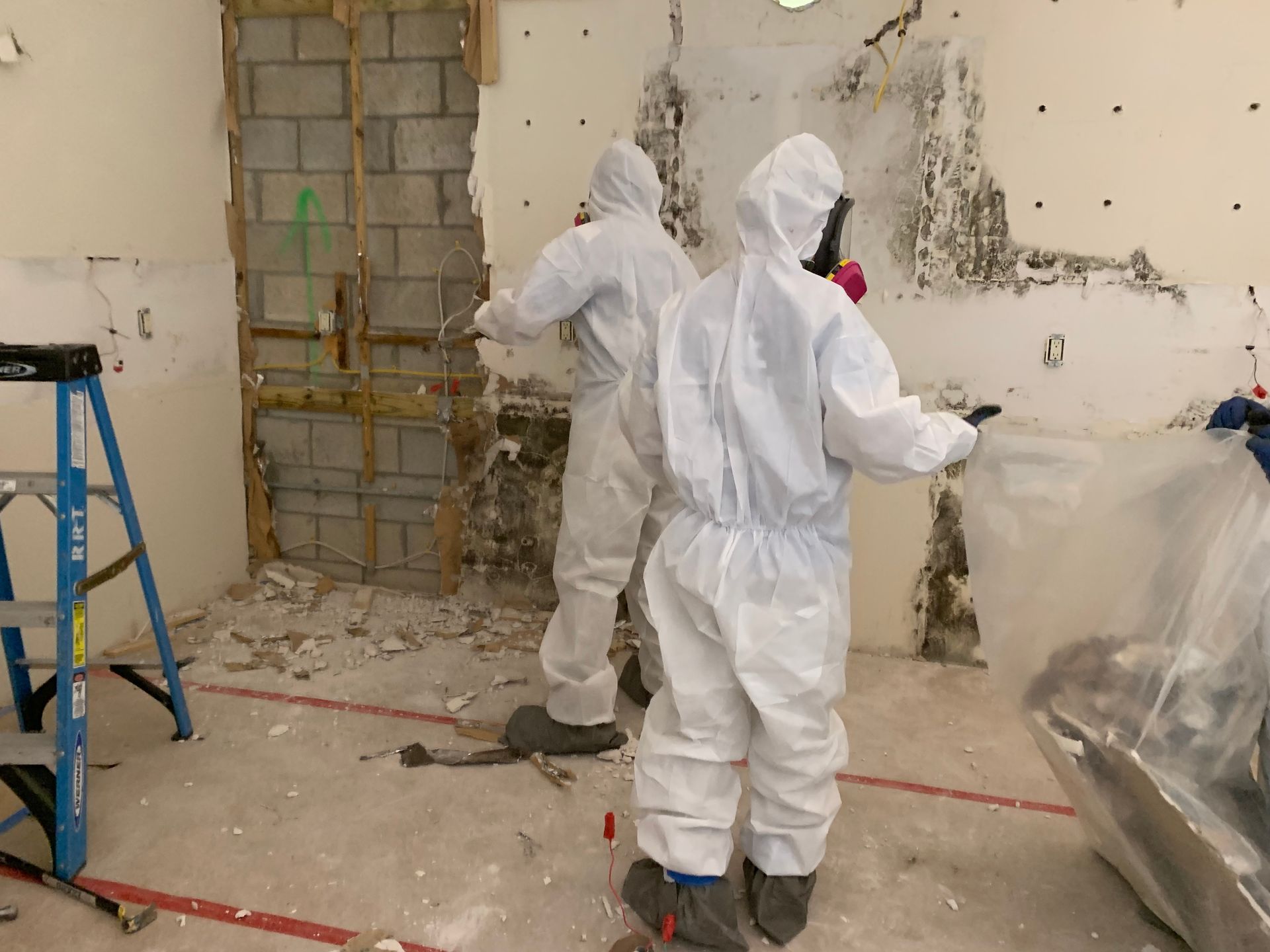 Two men in protective suits are working on a wall in a room.