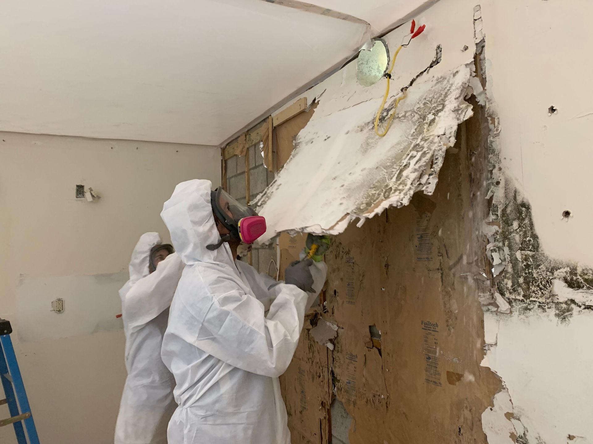 Two people in protective suits are working on a wall.