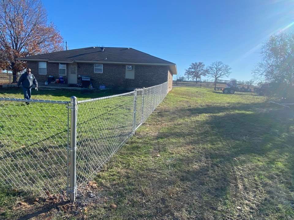A man is walking along a chain link fence in front of a house.
