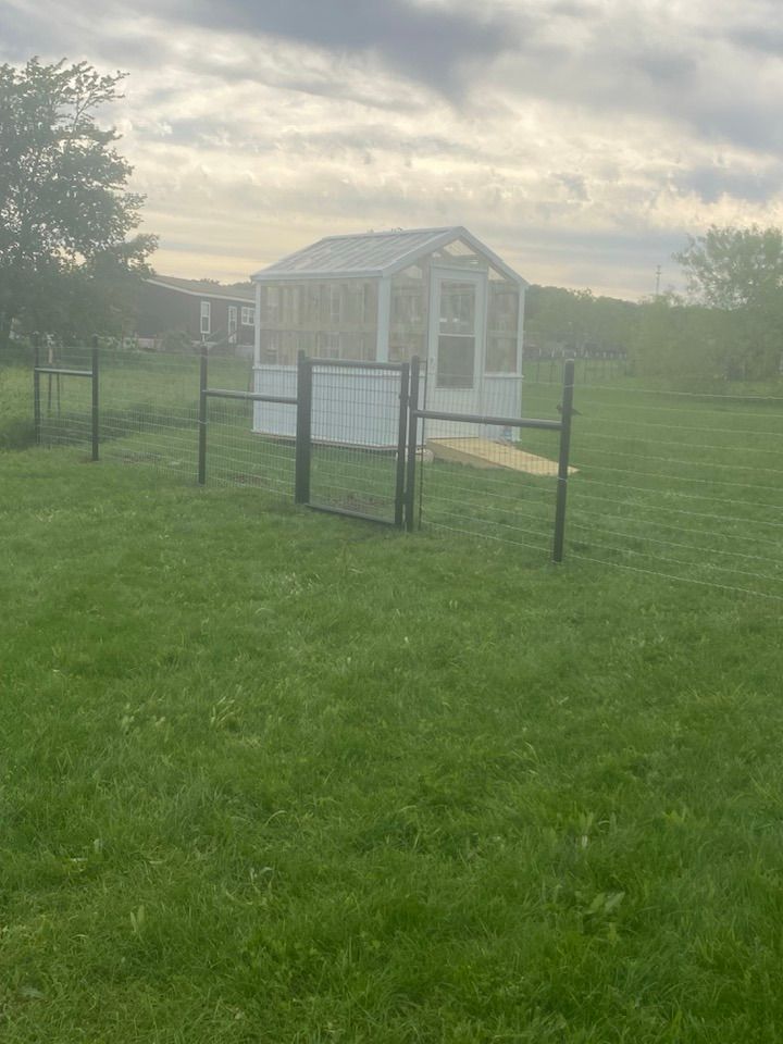 A greenhouse is sitting in the middle of a grassy field behind a fence.