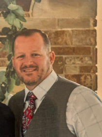 A man in a suit and tie is smiling in front of a brick wall.