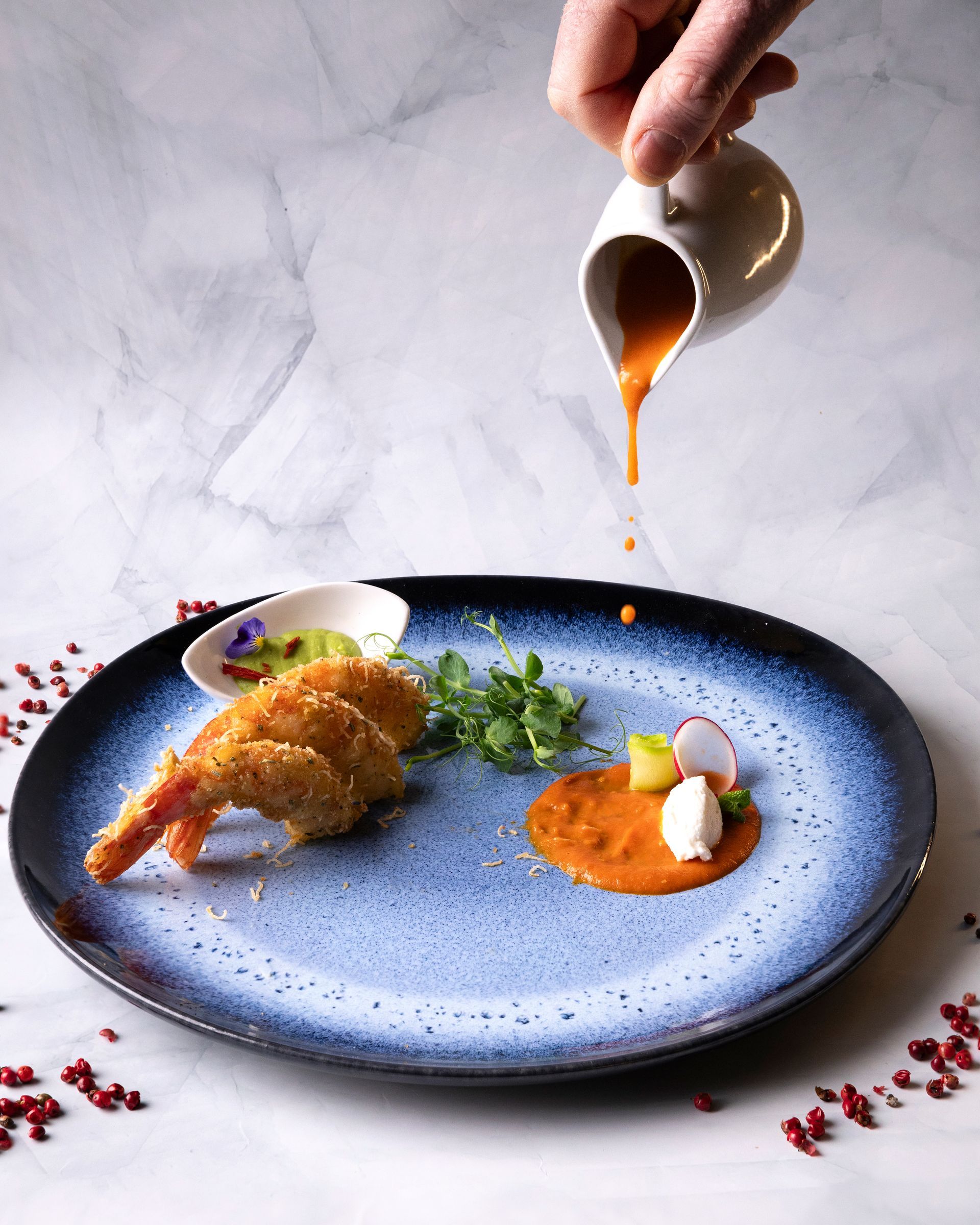 Un piatto di gamberi impanati con salsa versata da una piccola brocca. Altre guarnizioni includono una piccola salsa, verdure e ortaggi.