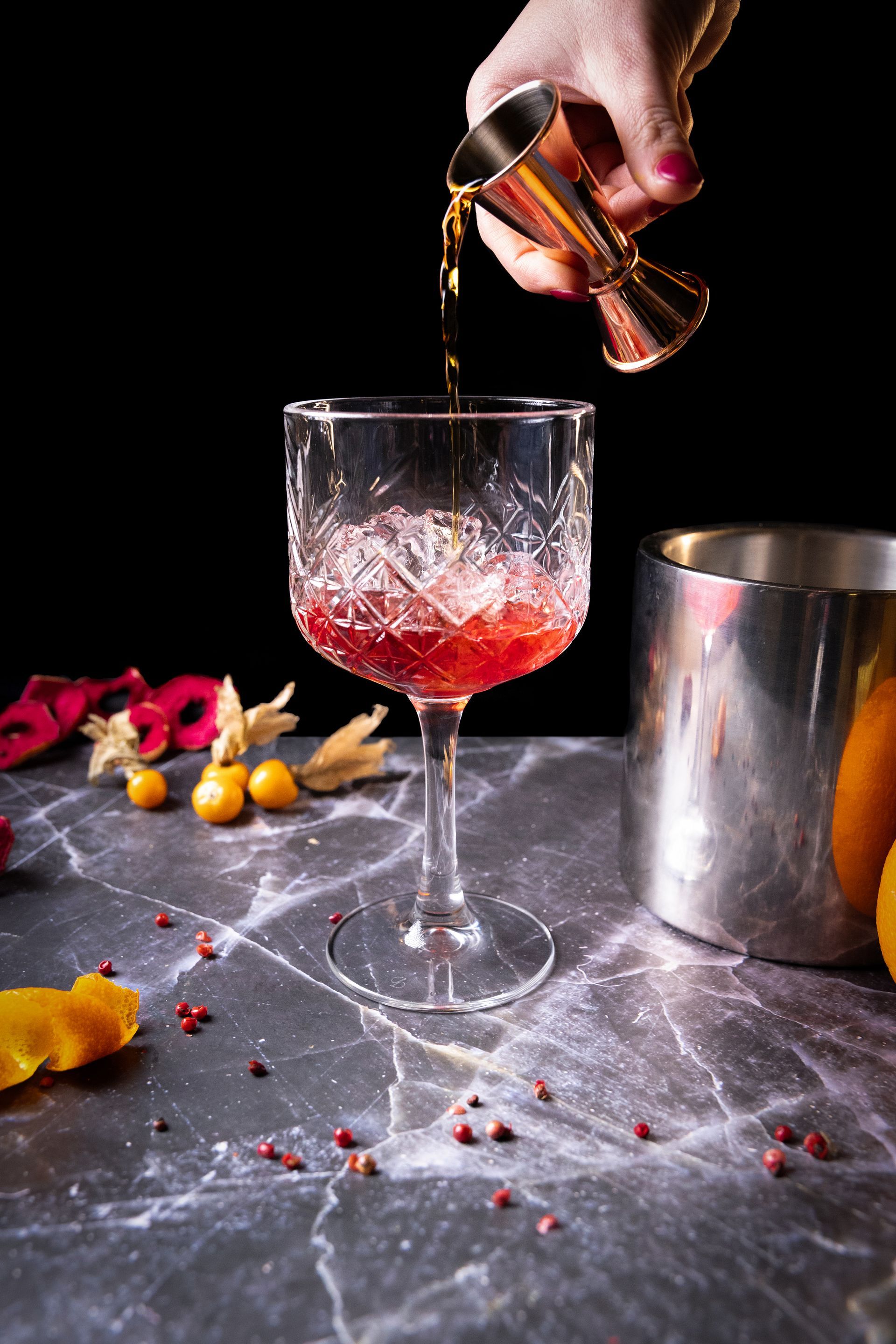 Cocktail versato da un misurino dorato in un bicchiere di cristallo, con uno shaker d'argento e guarnizioni su una superficie di marmo.