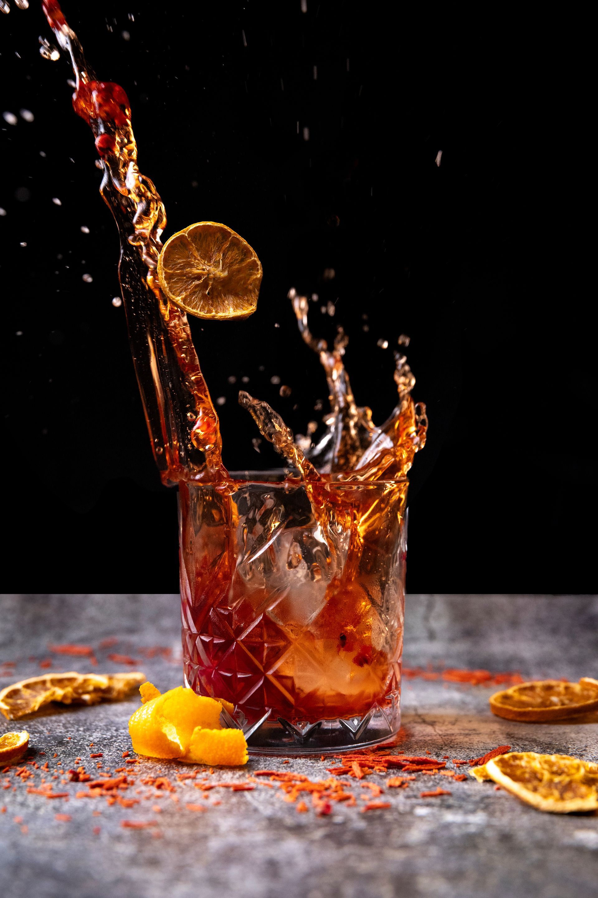 Un cocktail schizza fuori da un bicchiere di cristallo con ghiaccio, una fetta d'arancia essiccata sospesa e scorza d'arancia sulla superficie su uno sfondo scuro.