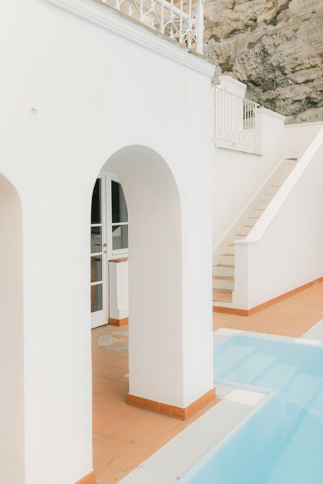 Un passaggio ad arco bianco conduce a una porta e a una scala che costeggia una piscina azzurra. L'edificio è addossato a una scogliera rocciosa.