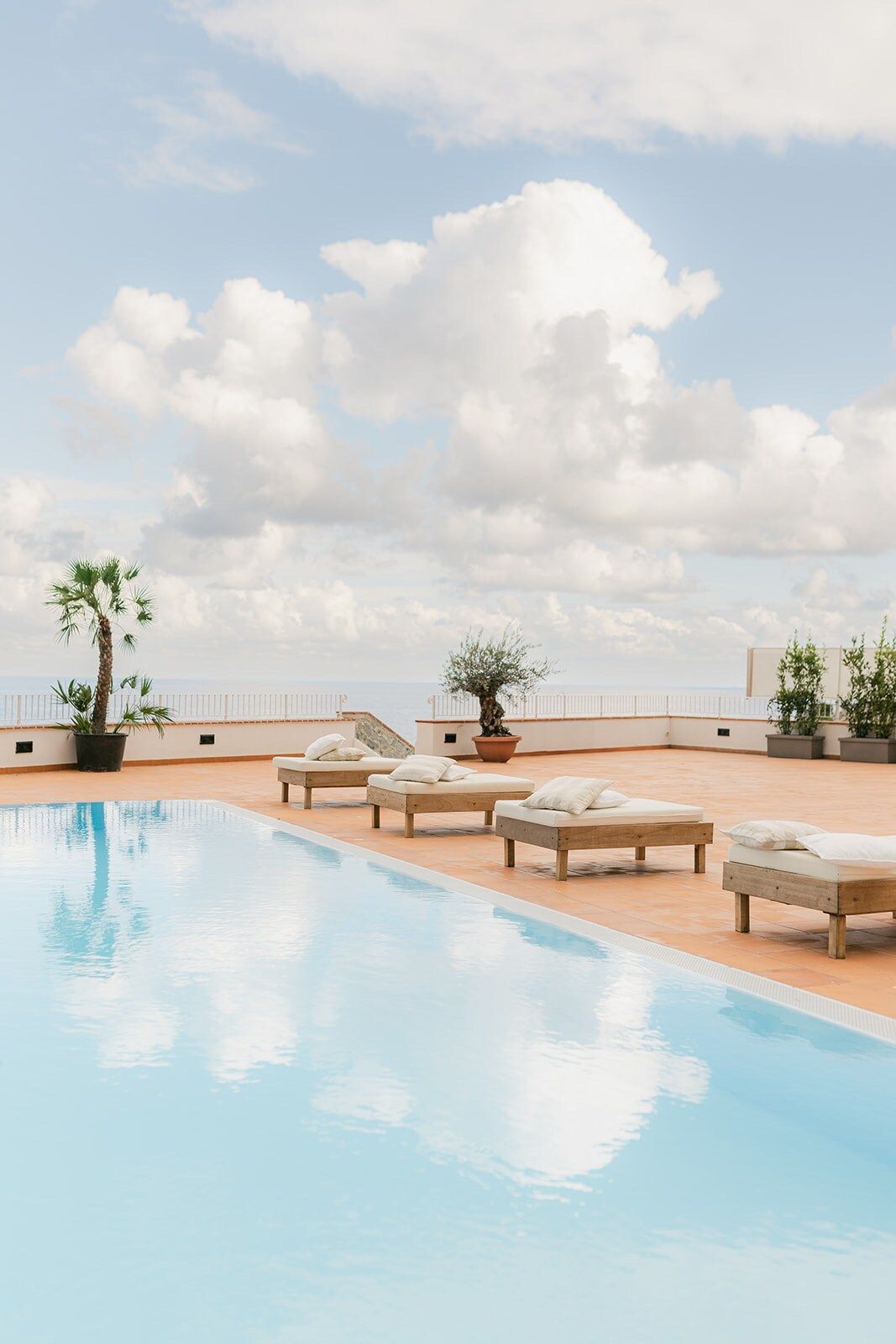Una piscina sul tetto con sedie a sdraio riflette il cielo, con alberi in vaso e vista sul mare nelle giornate di sole.