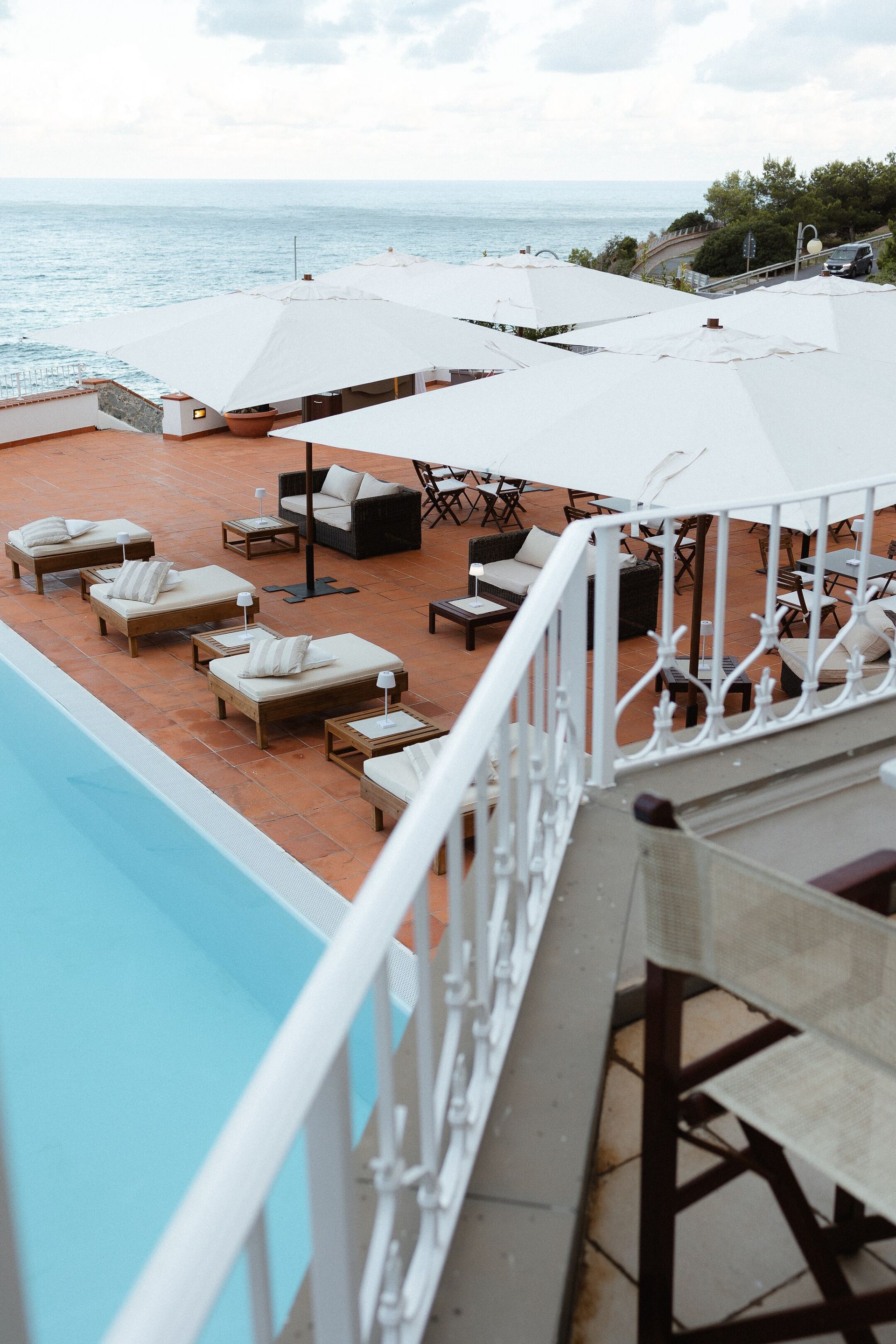 Un lounge sul mare con piscina blu, ombrelloni bianchi e sedute imbottite si affaccia sull'oceano. Una ringhiera bianca è in primo piano.