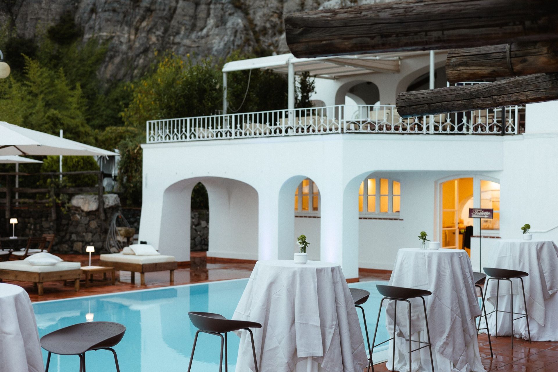 Bar all'aperto a bordo piscina con tavoli e sgabelli bianchi, un edificio bianco con archi e una montagna sullo sfondo.