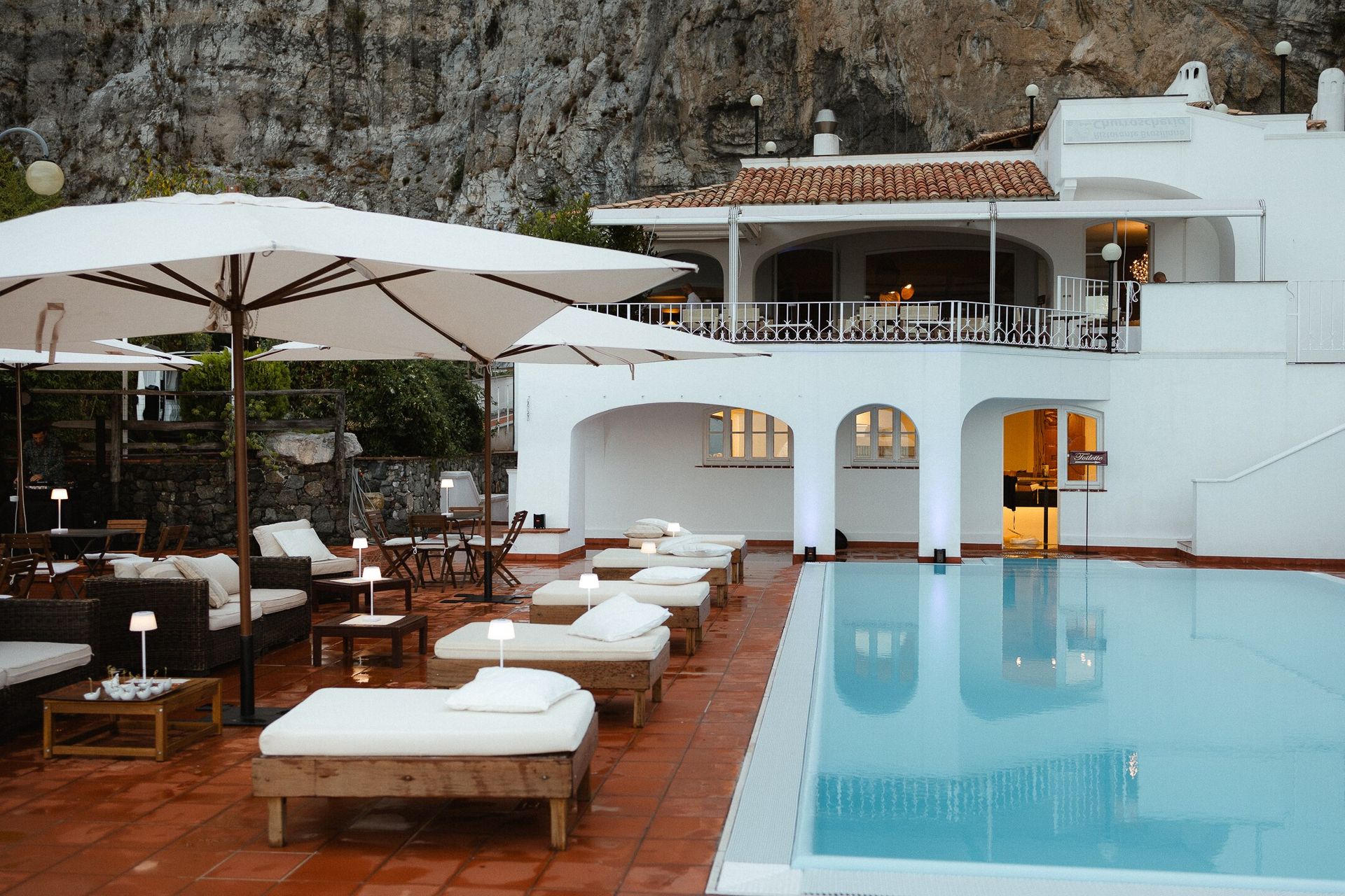 Scena di un resort a bordo piscina con sedie a sdraio, ombrelloni e un edificio bianco con balcone, vicino a una scogliera rocciosa.