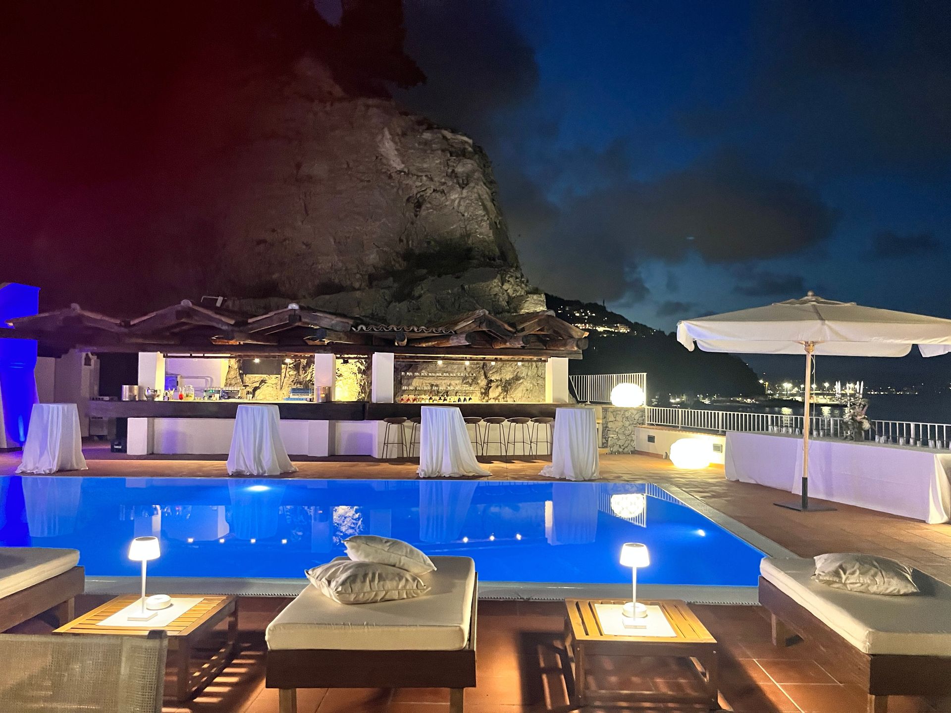 Bar a bordo piscina di notte con piscina illuminata, sedie a sdraio e una scogliera sullo sfondo.