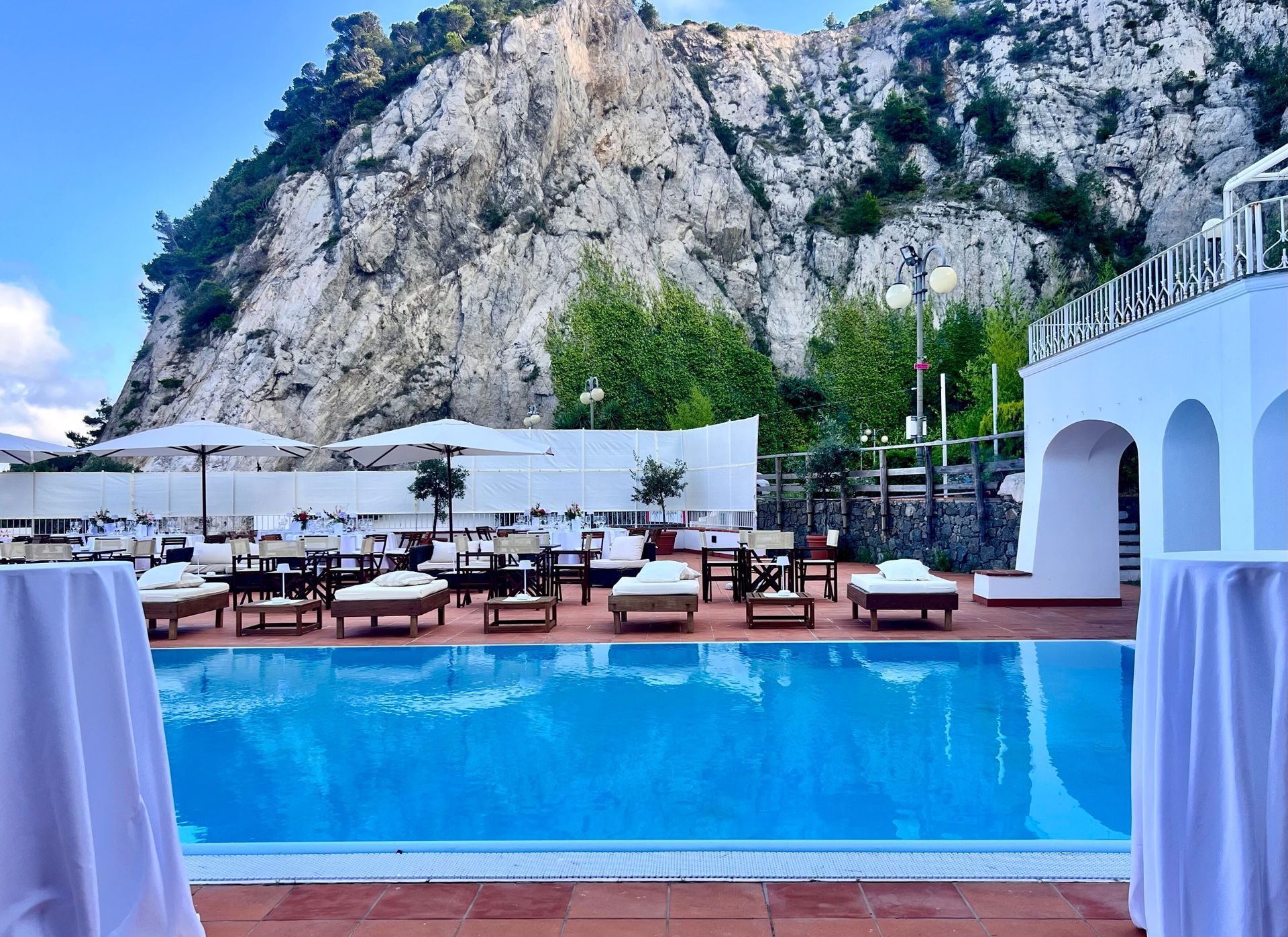 Una piscina con lettini e tavoli si affaccia su una montagna. Edifici bianchi e ombrelloni la circondano, sullo sfondo di un cielo azzurro.