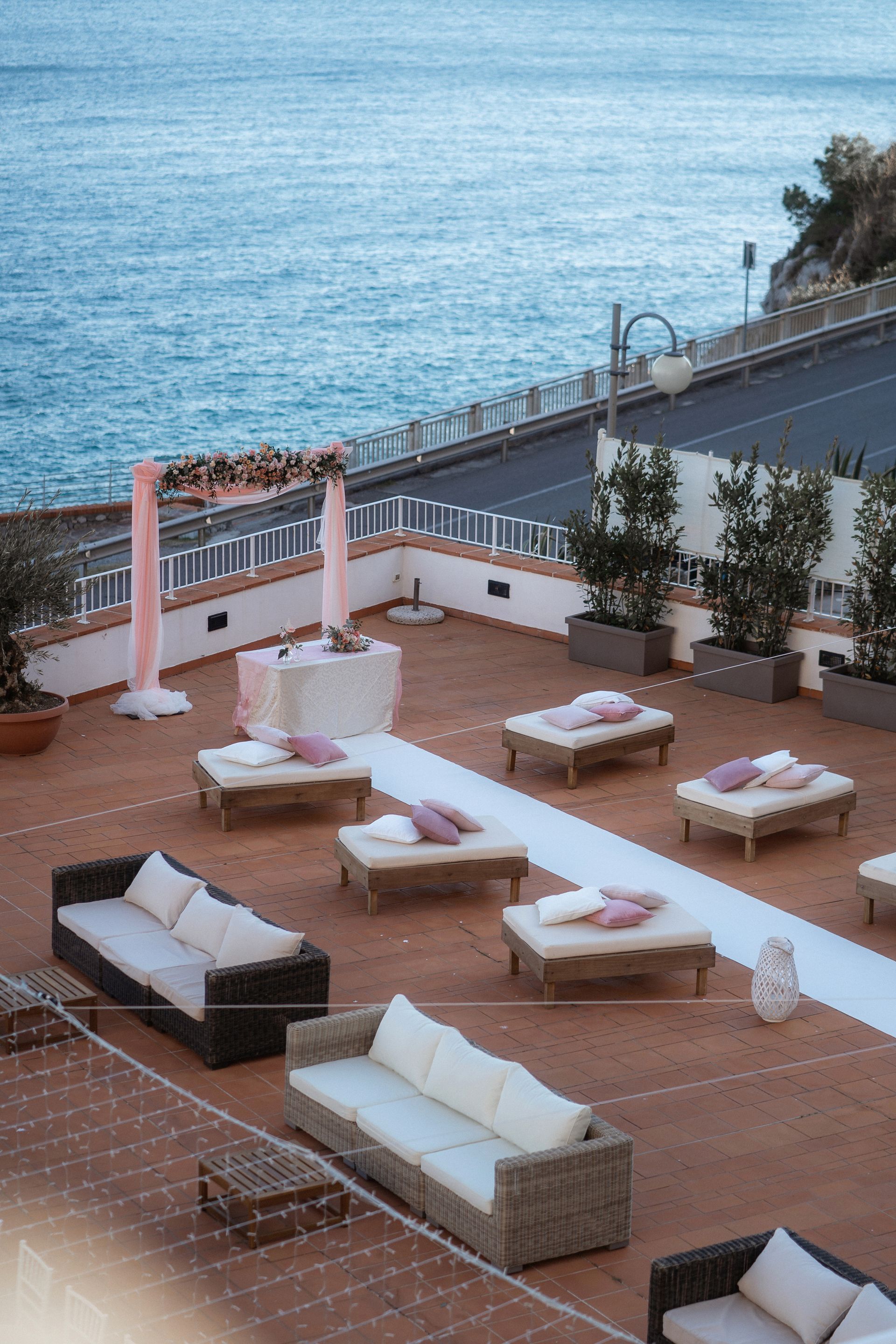 Location per matrimoni all'aperto con vista sul mare, dotata di divani, lettini e un arco floreale. Toni neutri e accenti rosa.