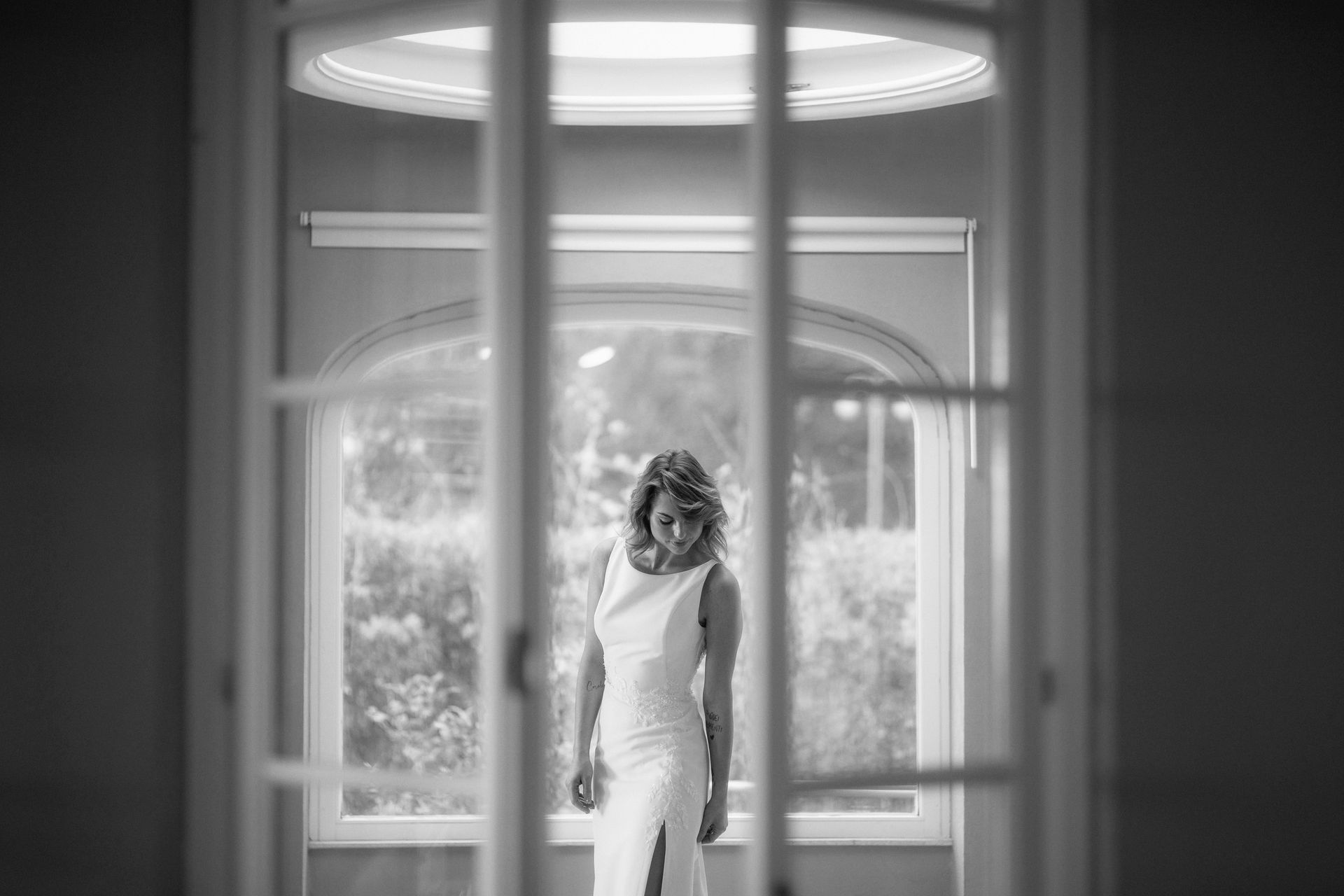 Una sposa in abito bianco è in piedi all'interno di un edificio, con lo sguardo rivolto verso il basso. È incorniciata da una grande finestra.