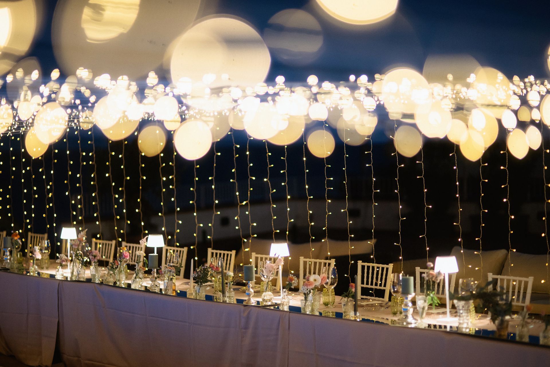 Lungo tavolo da banchetto apparecchiato per una festa serale, con luci a filo e lanterne luminose sopra la testa.