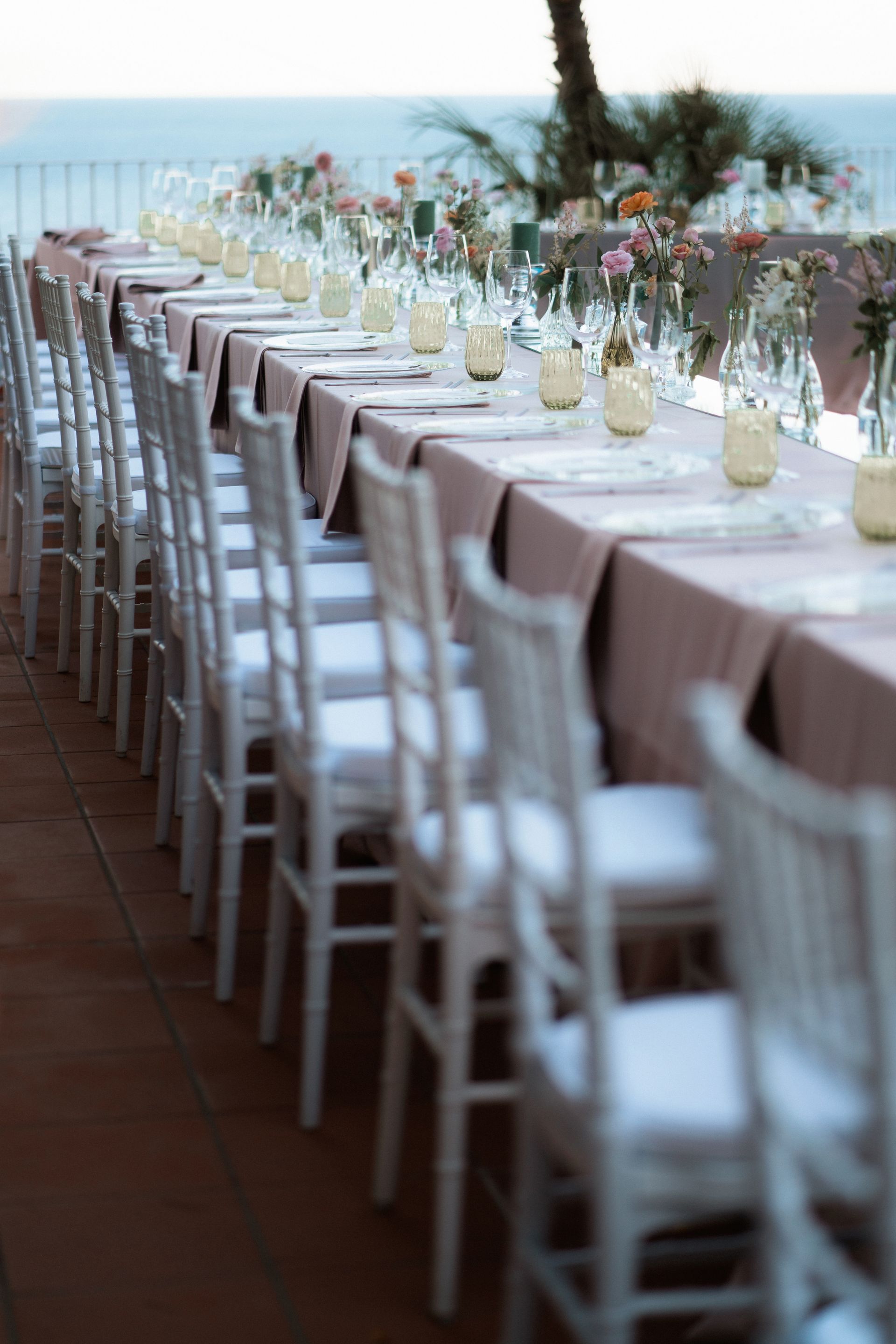 Lungo tavolo da ricevimento all'aperto, allestito con sedie bianche e centrotavola floreali, con vista sull'oceano.