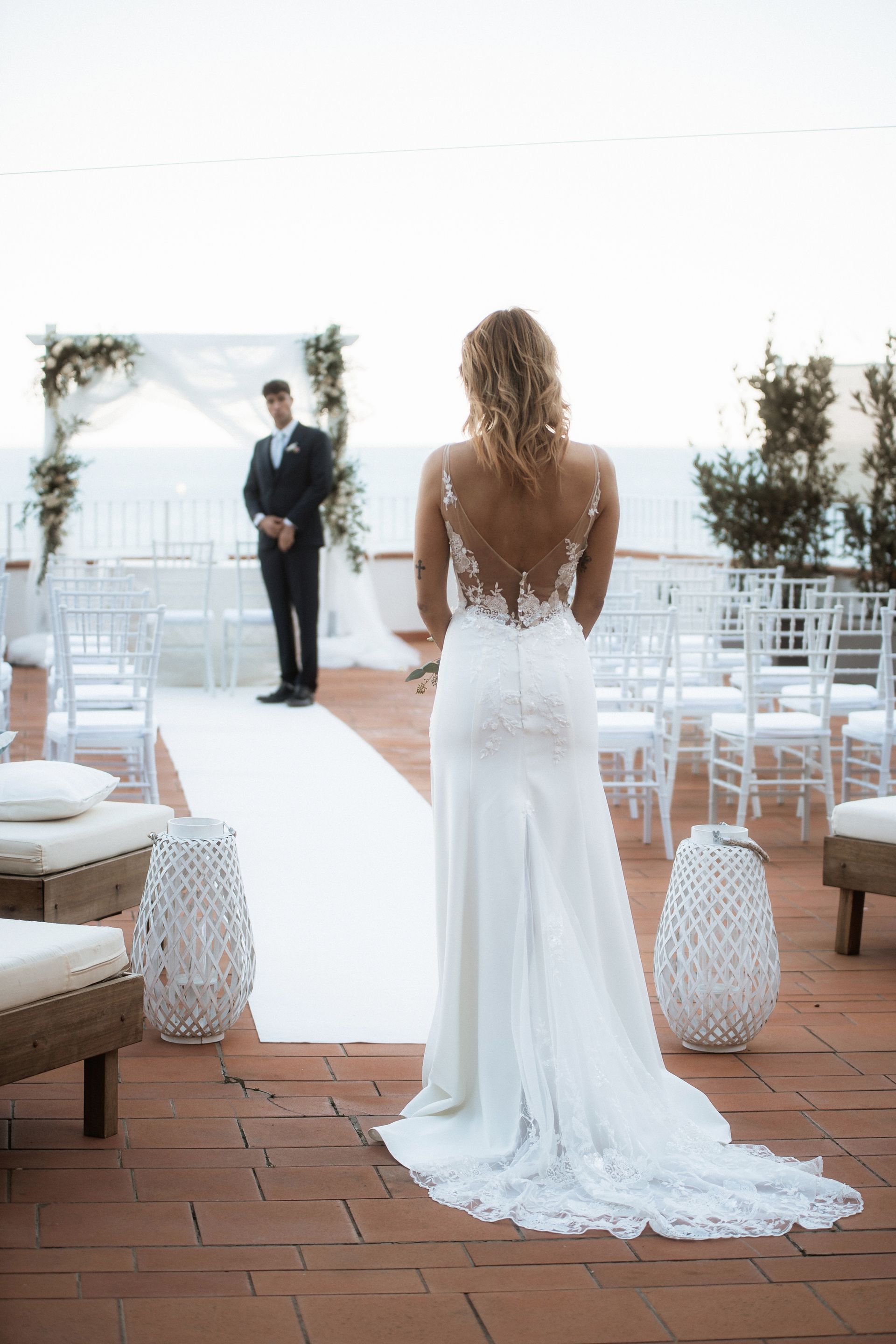 La sposa in un abito di pizzo con la schiena scoperta cammina verso lo sposo durante una cerimonia nuziale all'aperto. Sono visibili file di sedie e un arco decorato.
