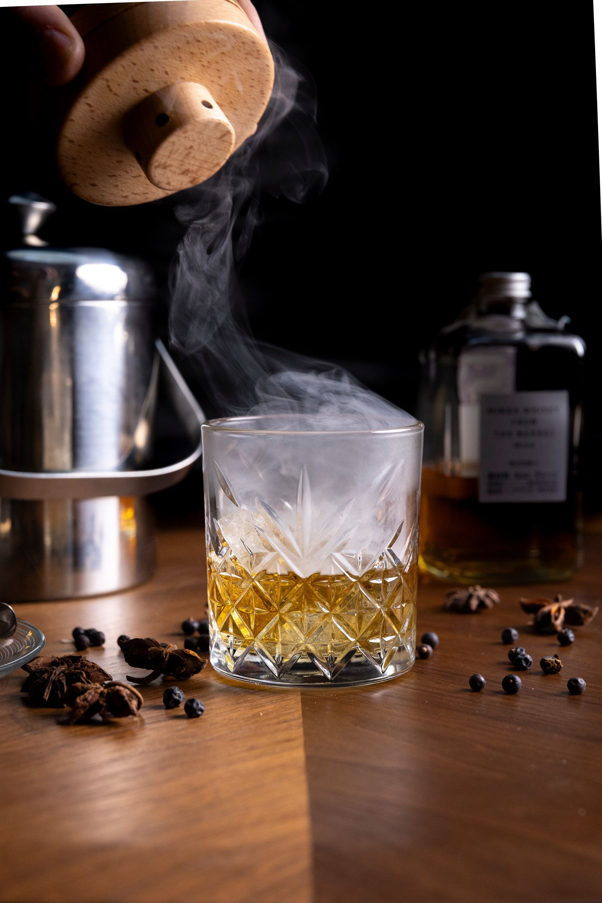 Cocktail affumicato: un bicchiere con liquido ambrato e fumo vorticoso, con un apparecchio per fumare, una bottiglia e spezie su una superficie di legno.