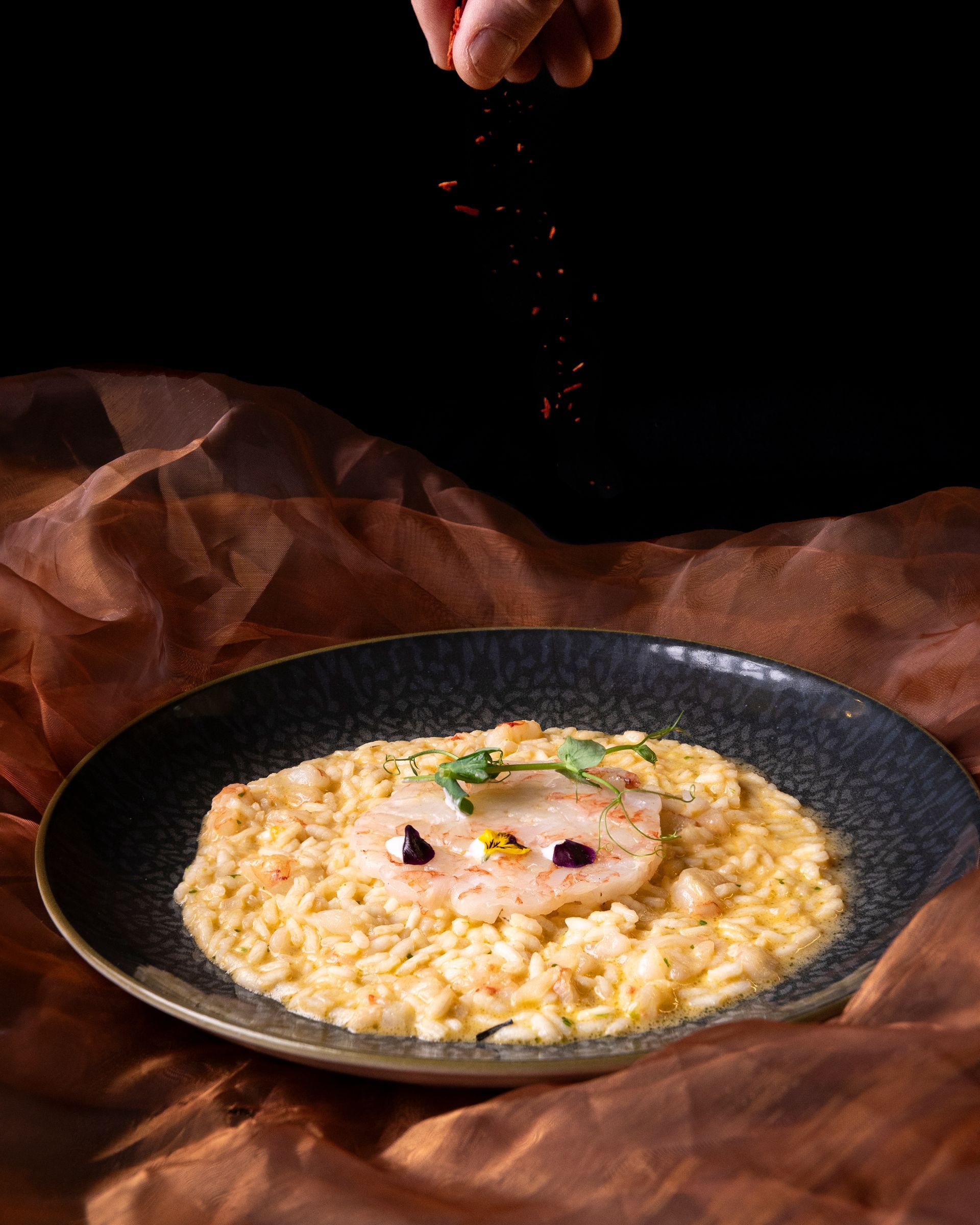 Un piatto di risotto con pesce scottato e cosparso di spezie rosse. Sfondo scuro con tessuto marrone.