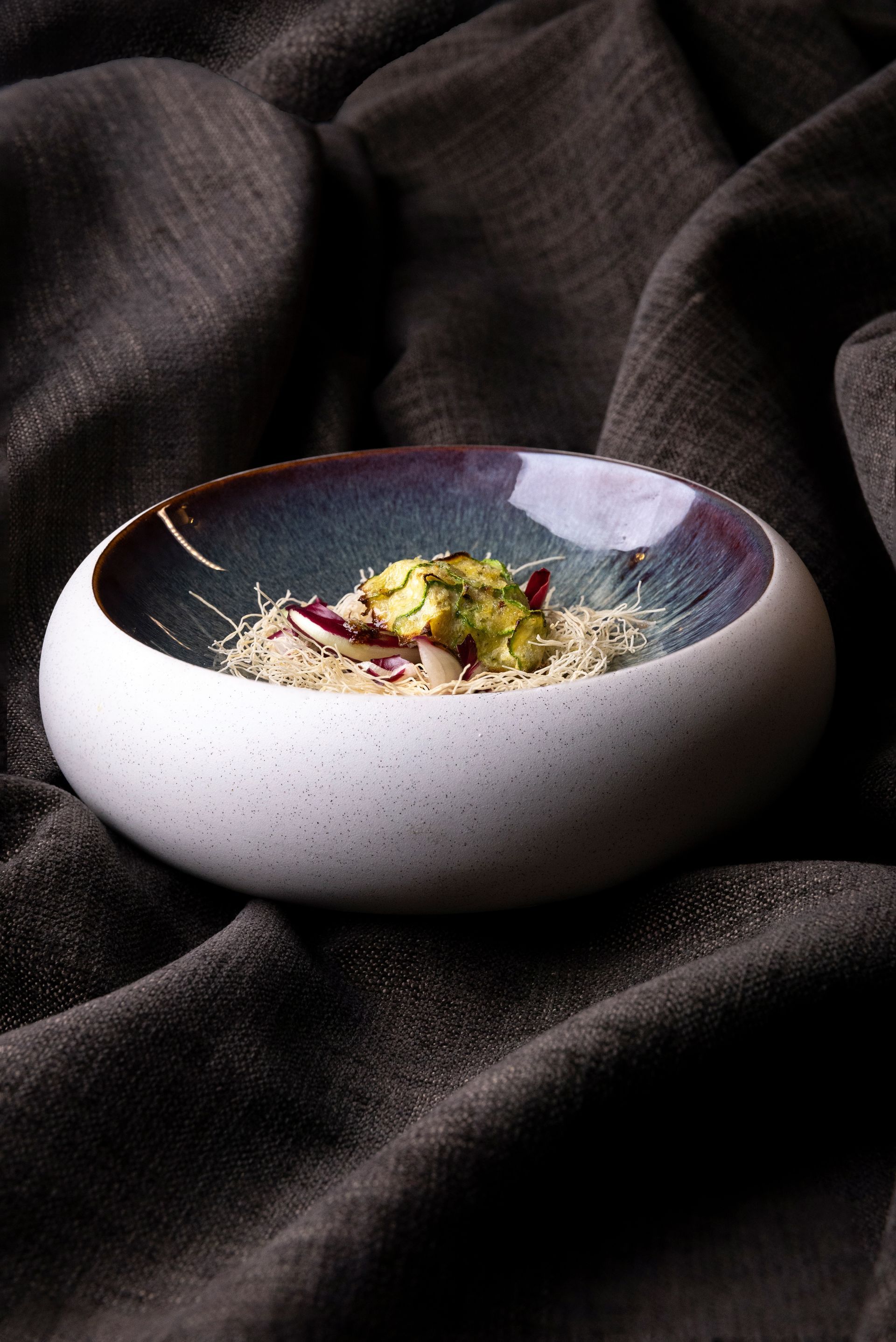 Una ciotola bassa, bianca e punteggiata, con un bordo viola-bluastro, contiene una piccola porzione di cibo su un tessuto grigio scuro testurizzato.