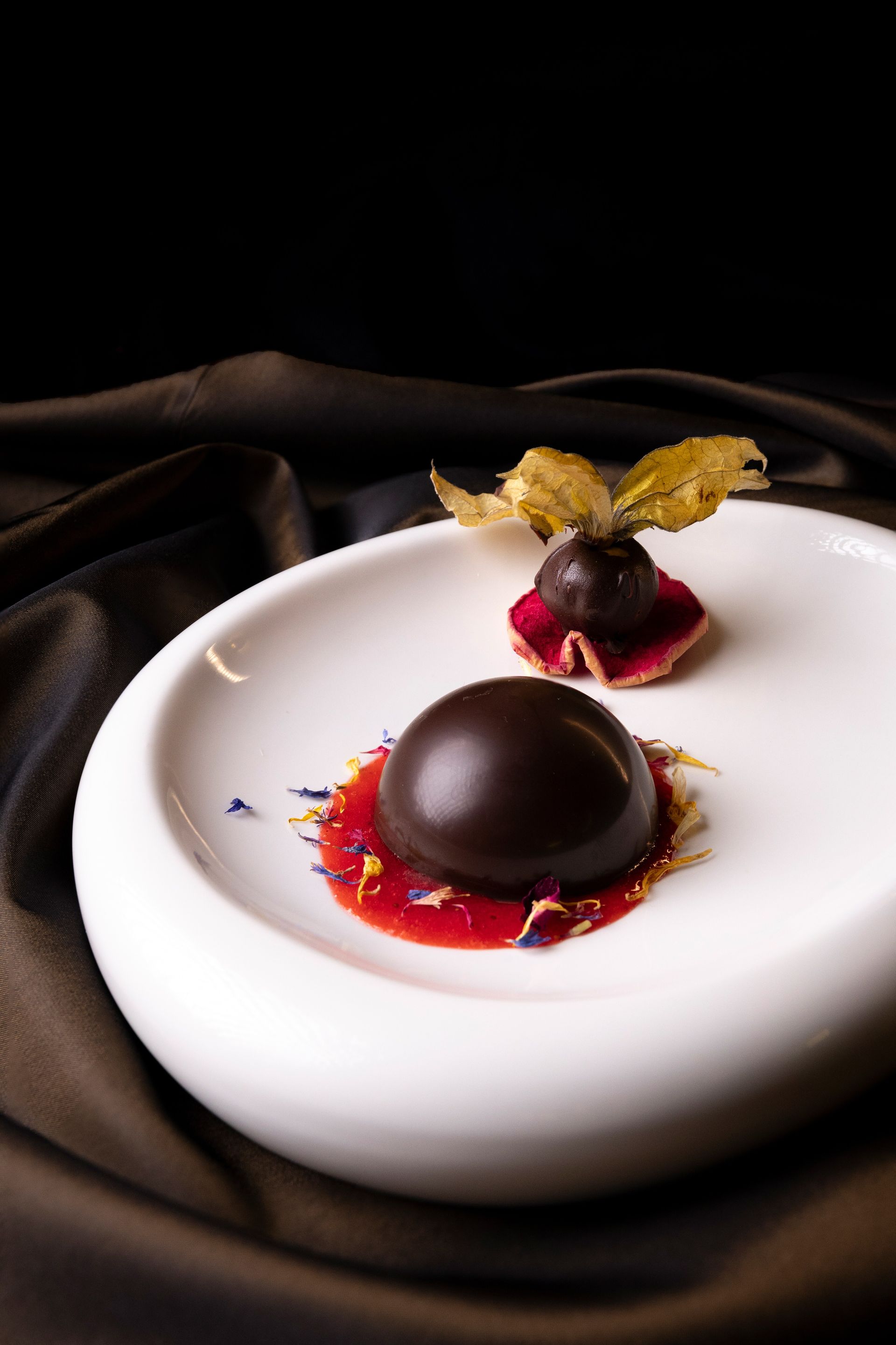 Un dessert al cioccolato fondente su un piatto bianco, guarnito con una piccola bacca e petali di fiori, adagiato su un panno scuro.