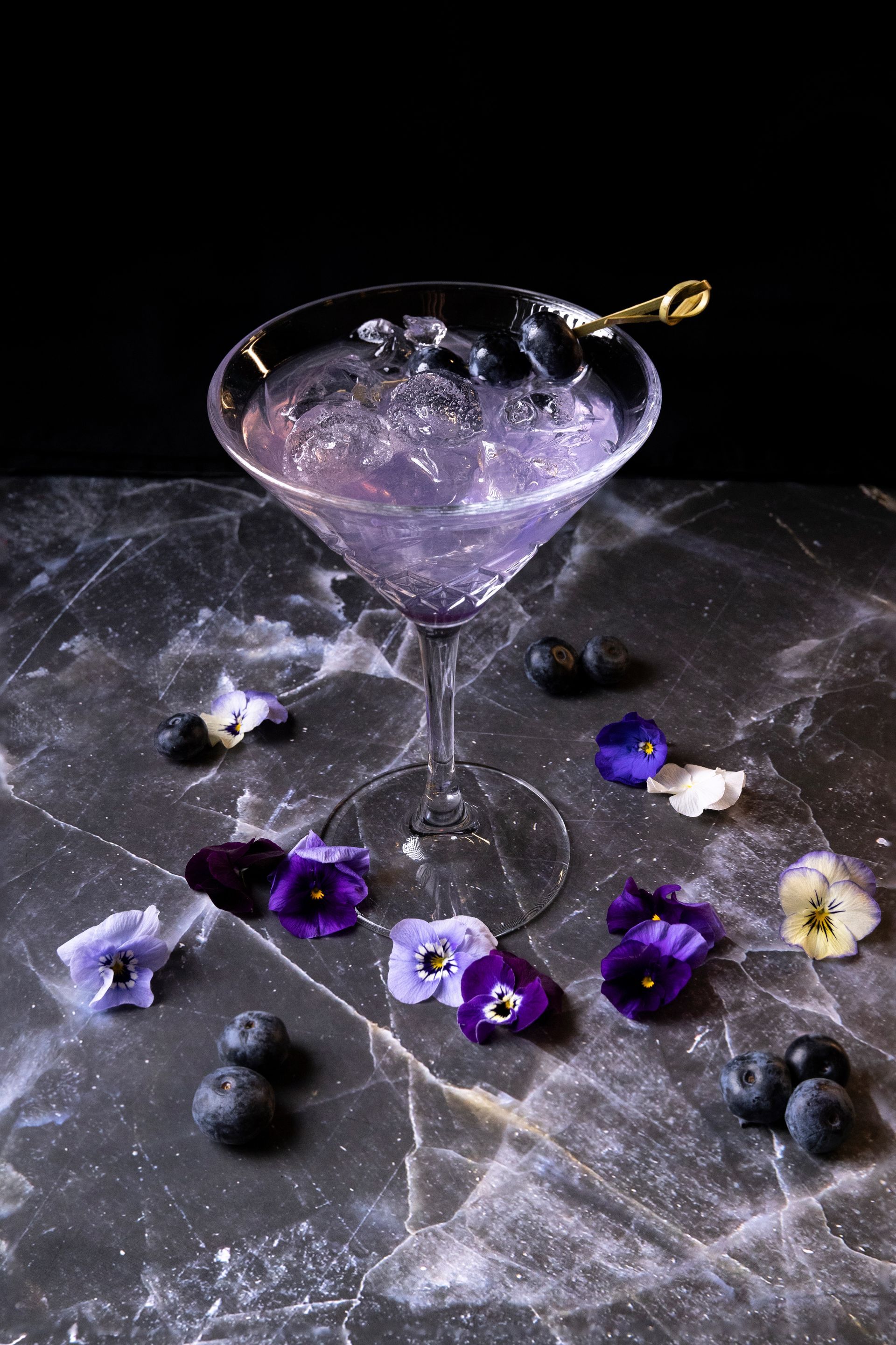 Un cocktail alla lavanda in un bicchiere da martini su una superficie di marmo, decorato con mirtilli e viole del pensiero colorate.
