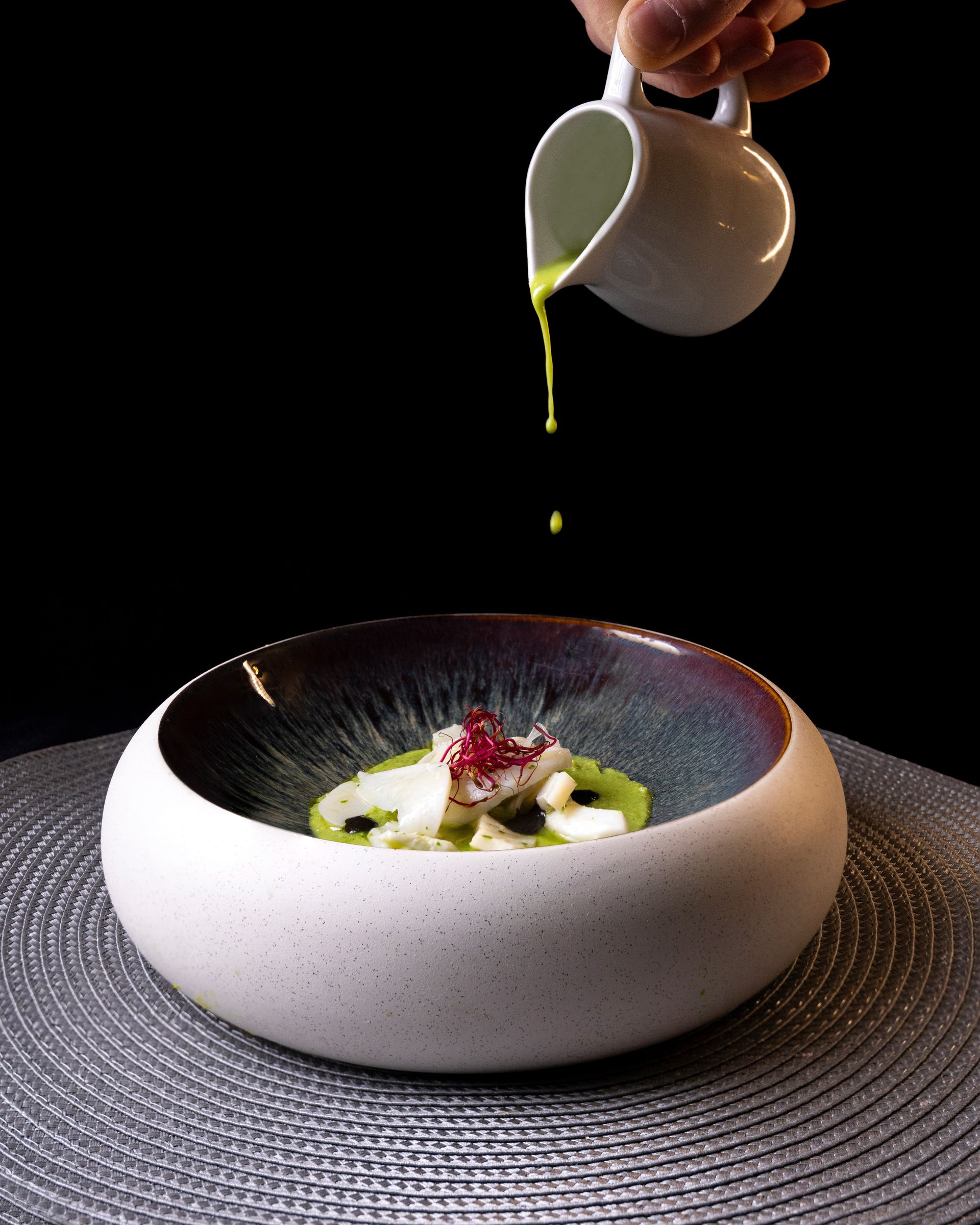 Mano che versa la salsa verde su una ciotola bianca contenente cibo. La ciotola ha un bordo blu scuro ed è appoggiata su un tappetino ruvido.